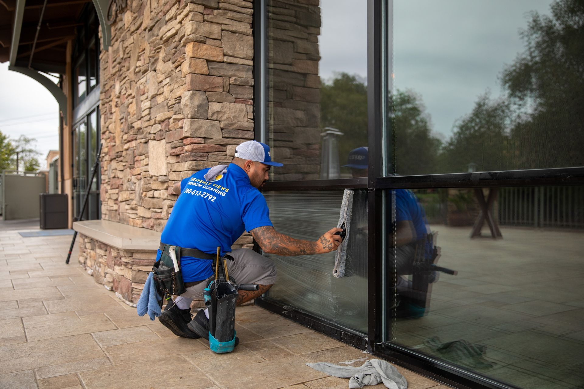 Worker in blue uniform cleaning a glass storefront window beside a stone building.