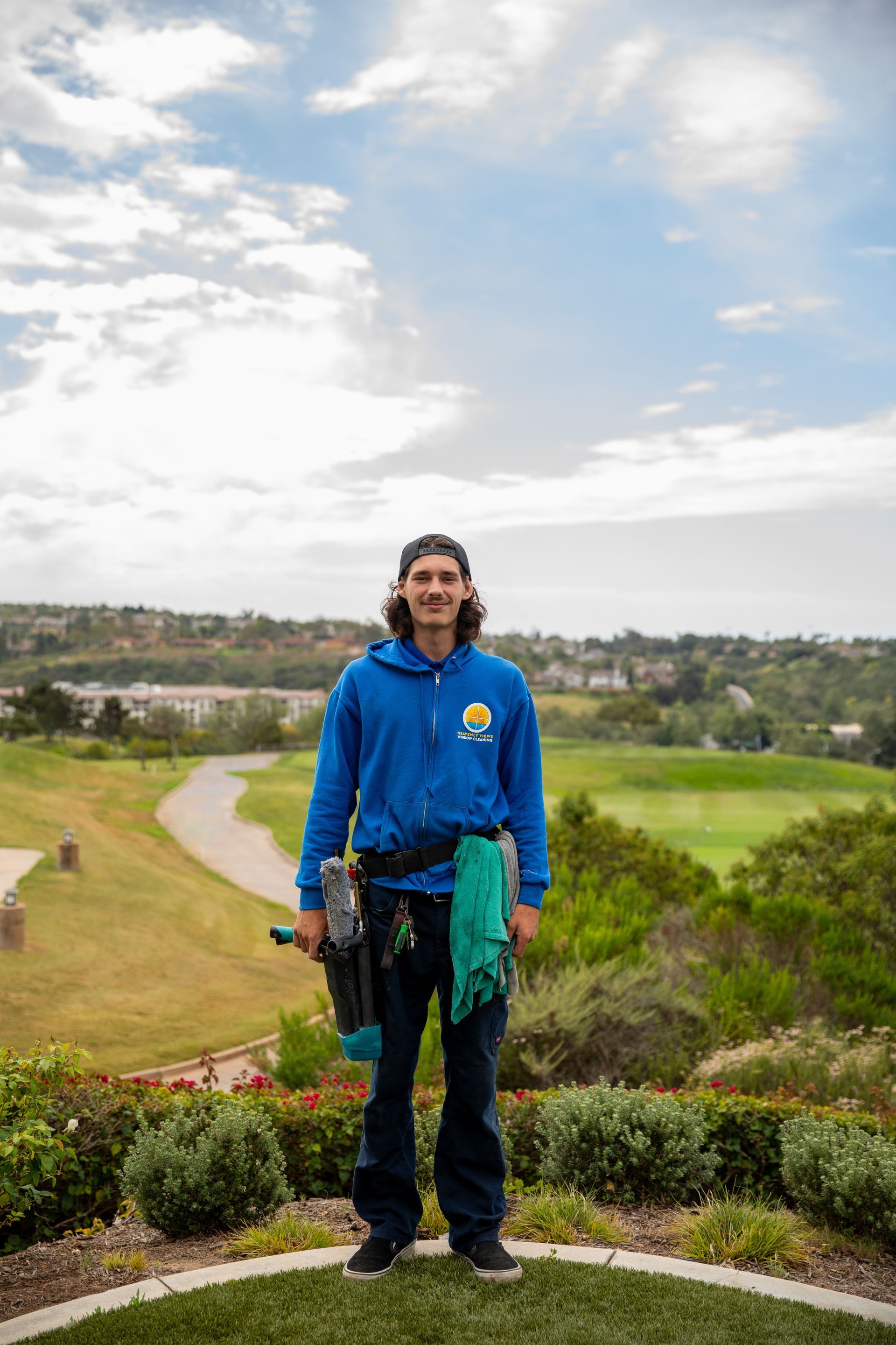 Person in blue hoodie standing on a scenic hillside path with fields and cloudy sky in the background