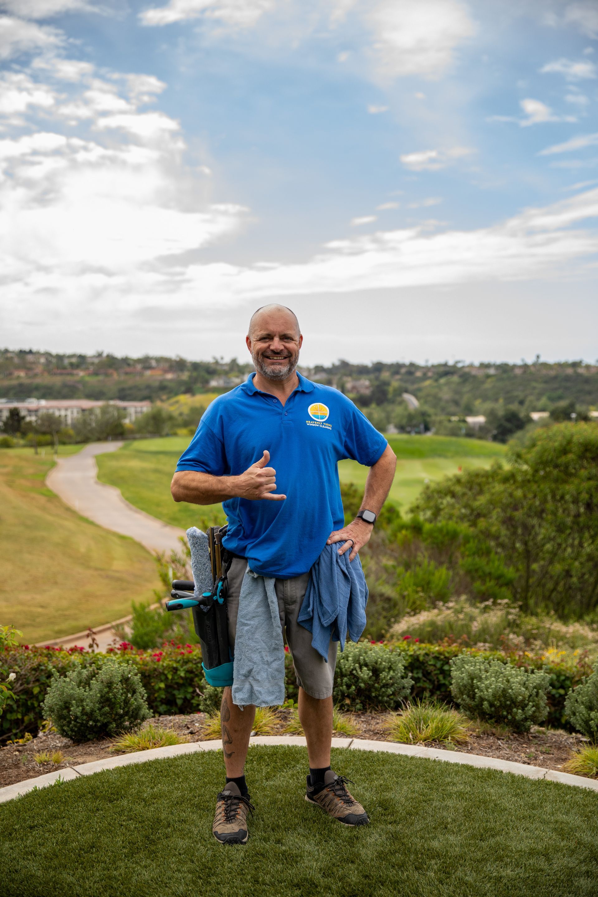 Man in blue shirt and shorts giving thumbs-up on a grassy hill overlooking a park and winding path