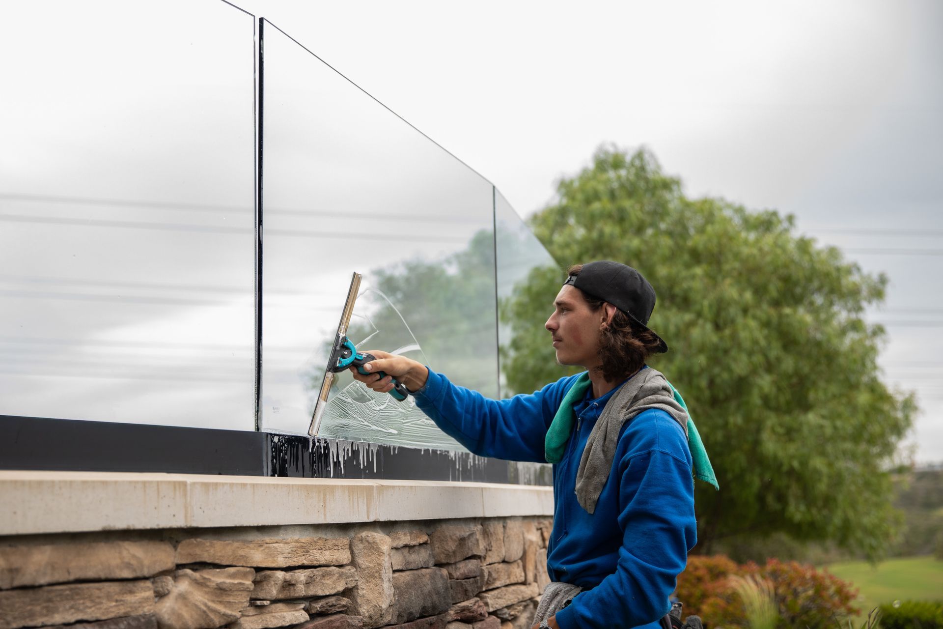 Worker cleaning a glass panel outdoors with a squeegee near a stone wall and trees