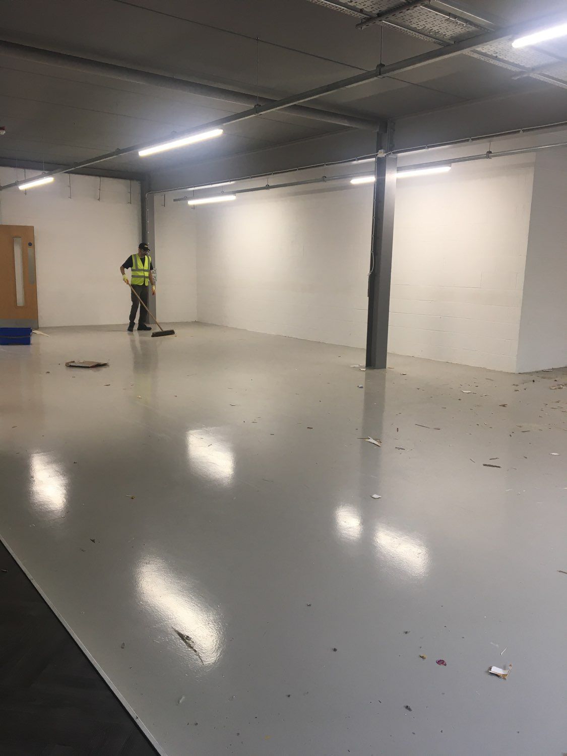 Empty warehouse-like room with glossy floor, white walls, and a worker in a yellow safety vest standing near the back.