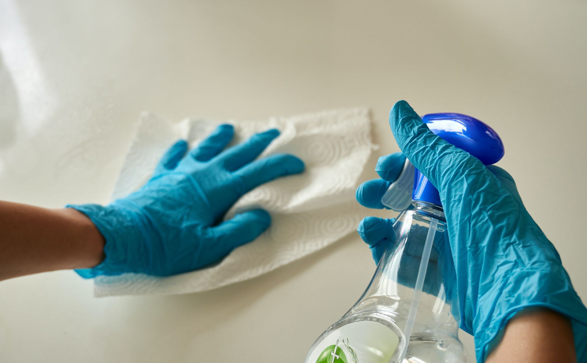 Gloved hands wiping a white surface with a cloth and spraying cleaner from a bottle