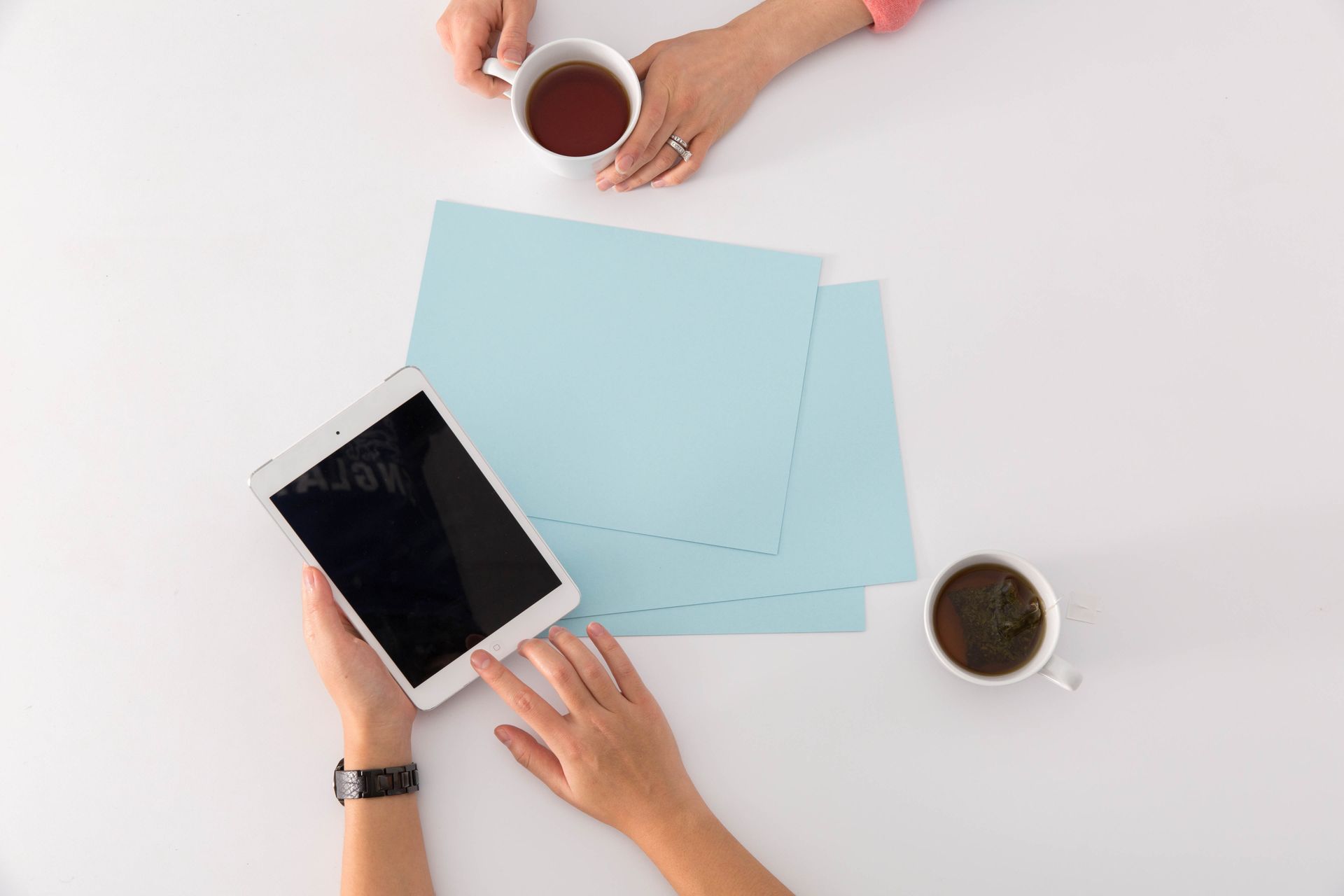 Overhead view of hands with a tablet, blue papers, and two coffee cups on a white desk