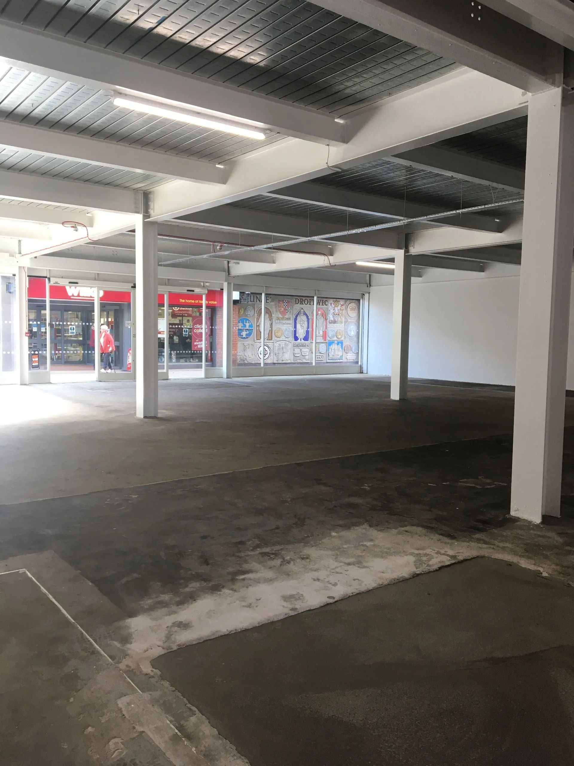 Empty covered parking area with white columns, dark pavement, and storefronts in the background