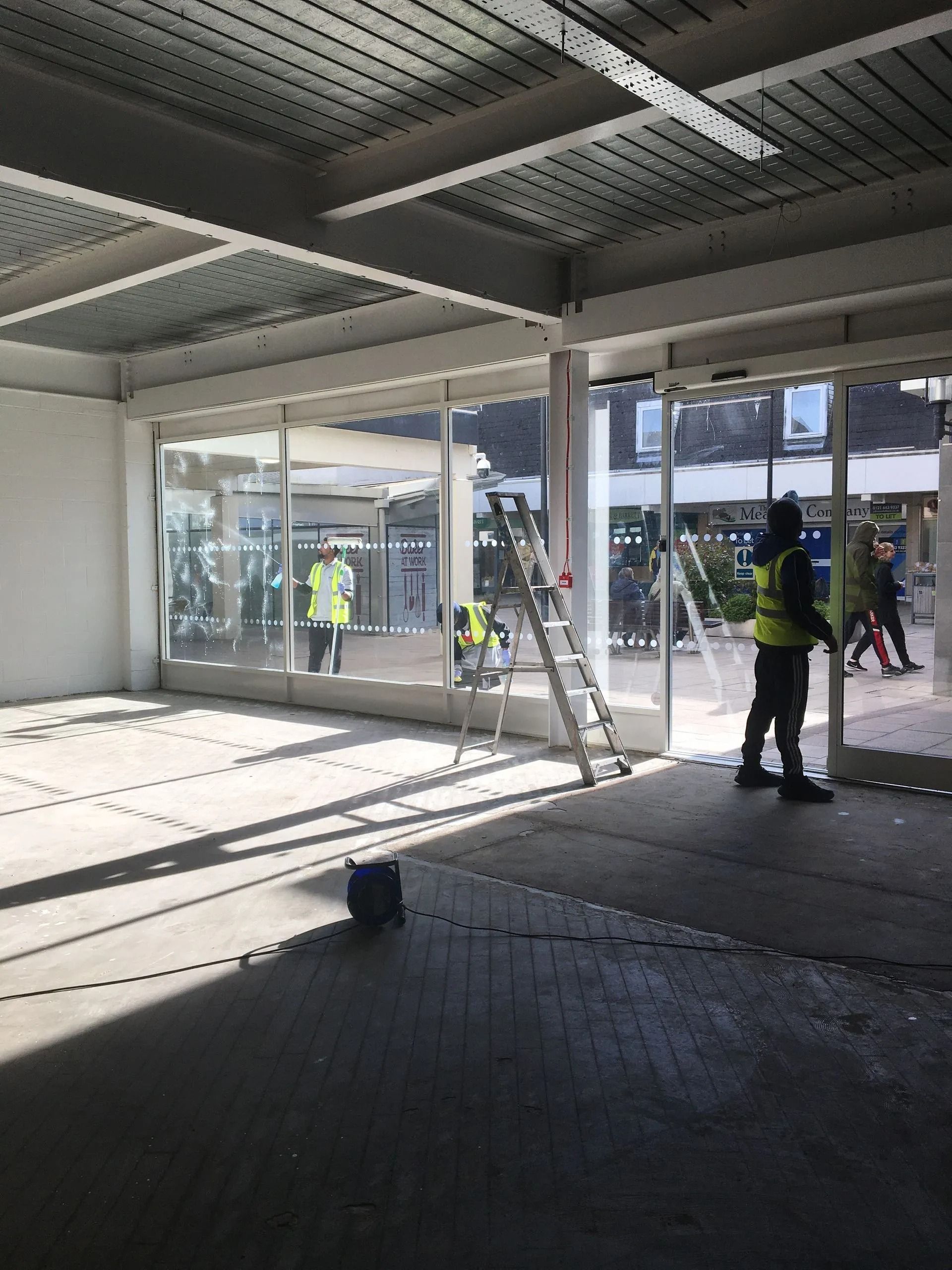 Bright indoor lobby with large glass walls and several workers in high-visibility vests outside.