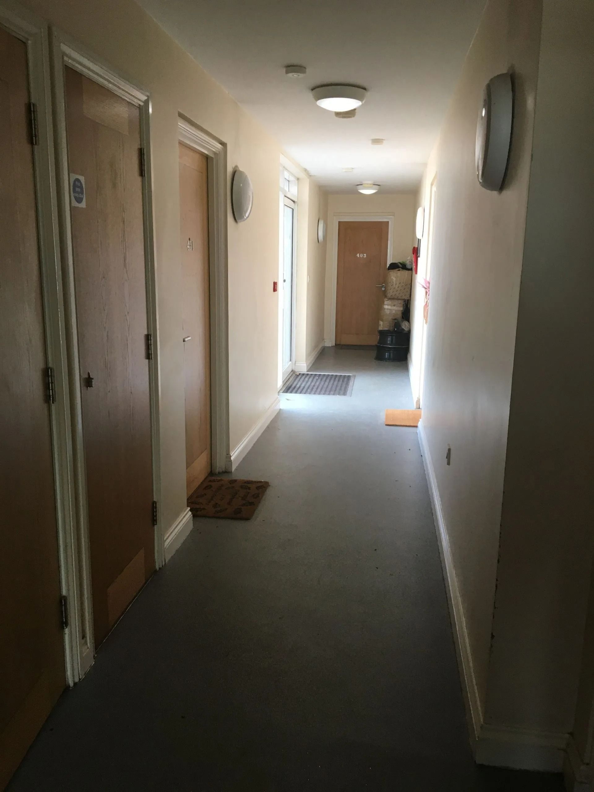 Narrow apartment hallway with beige walls, carpet, several doors, and a bright door at the end.