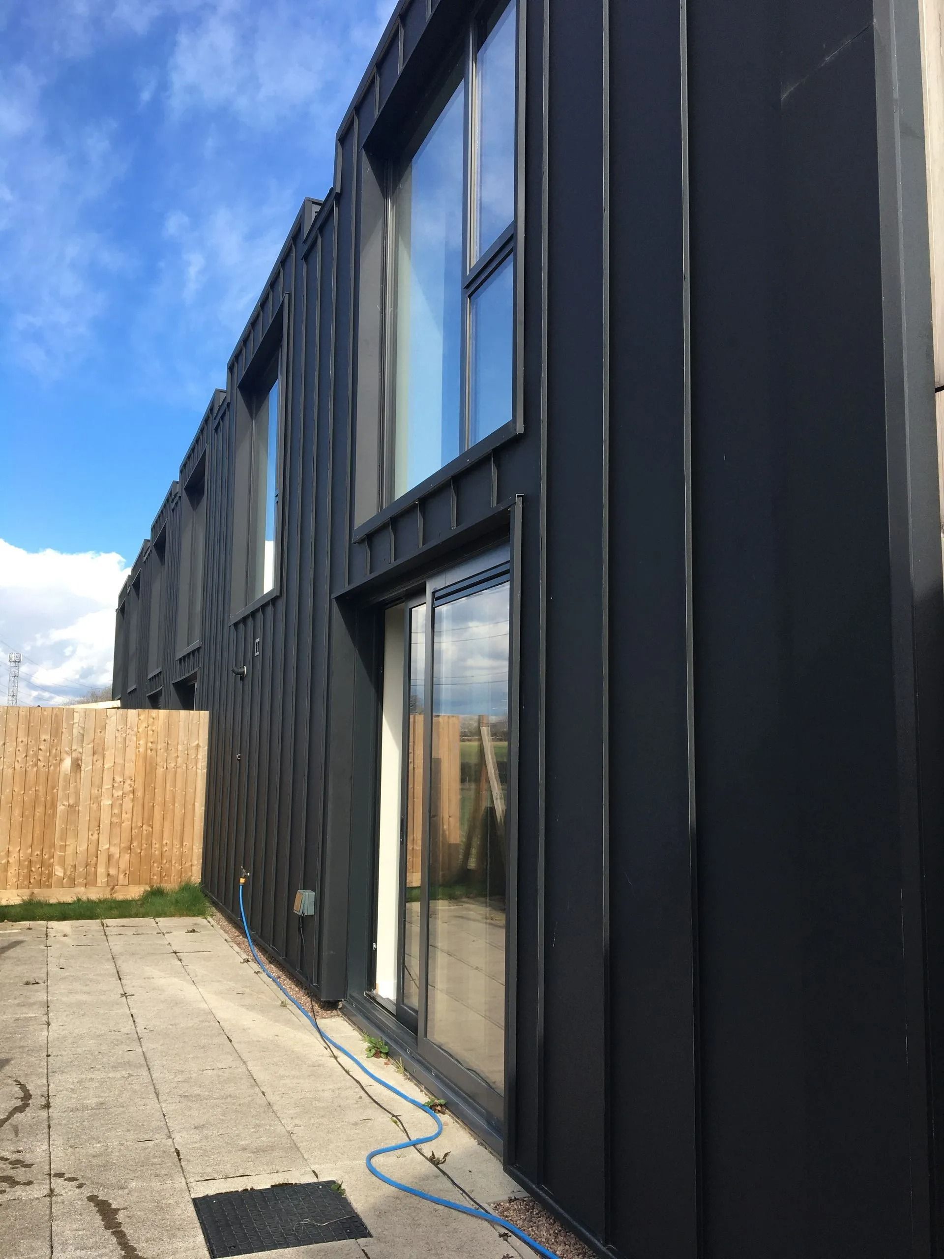 Black modern house exterior with tall windows and sliding glass door beside a paved yard and fence under blue sky