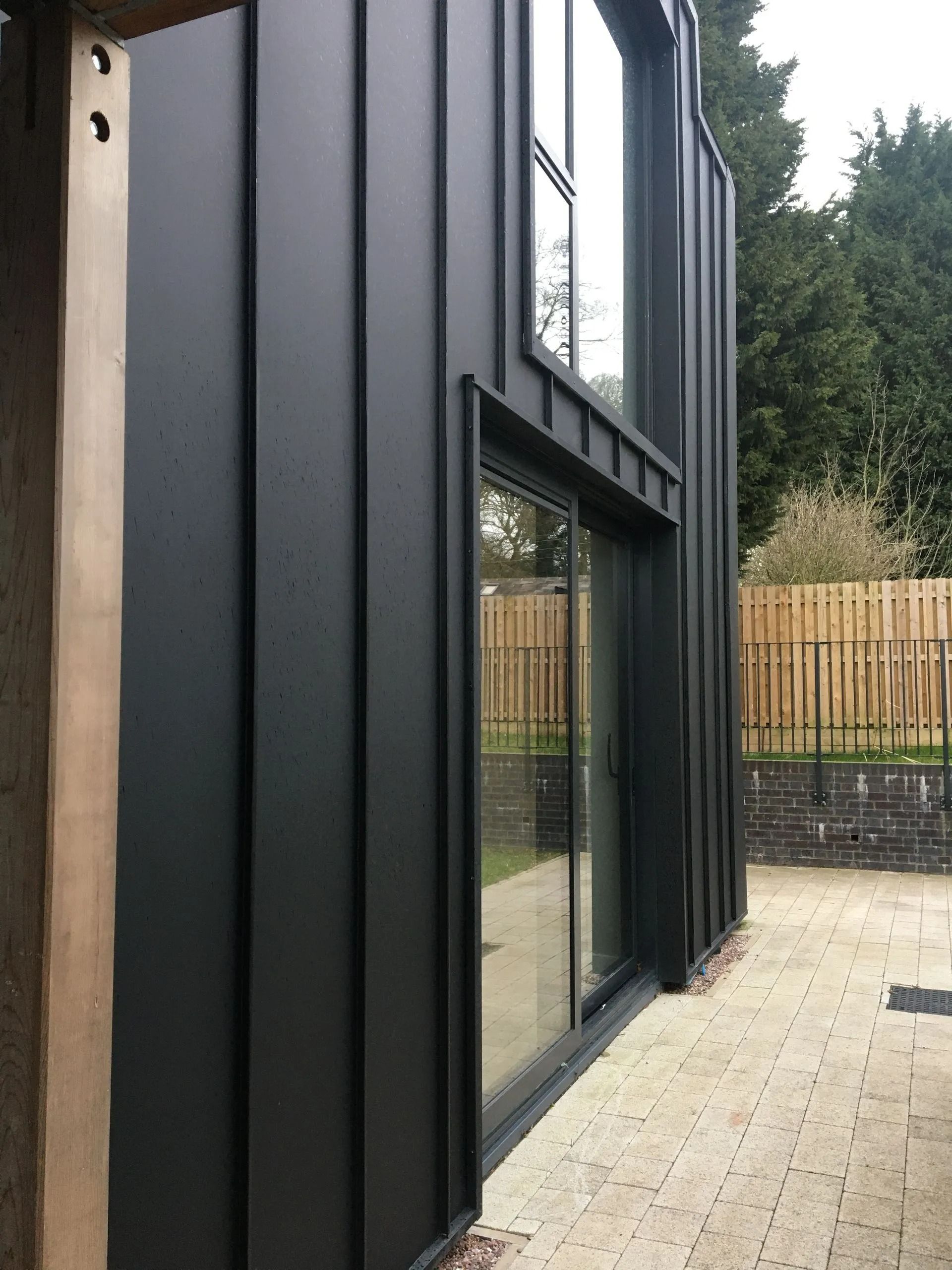 Black modern building exterior with large windows beside a paved yard and wooden fence