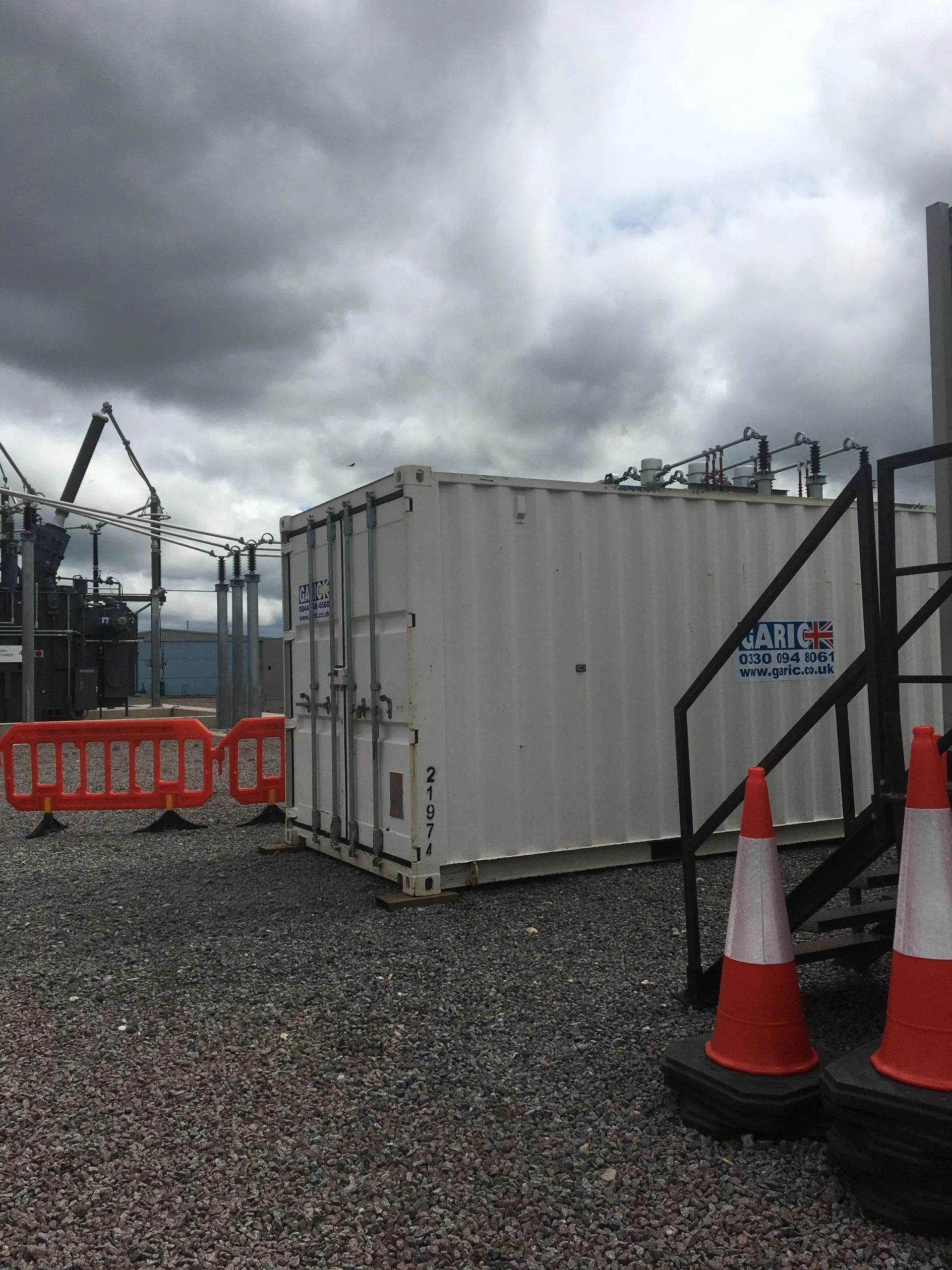 Industrial yard with white shipping container, orange barriers, traffic cones, and cloudy sky