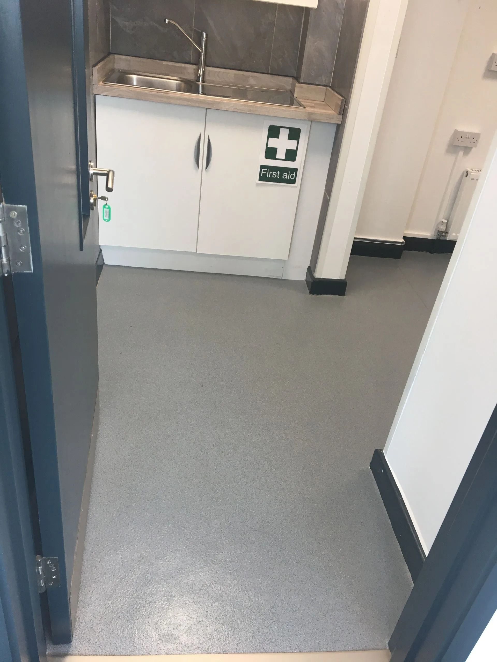 Empty hallway with white cabinets, gray floor, and a first-aid cross sign on the wall.
