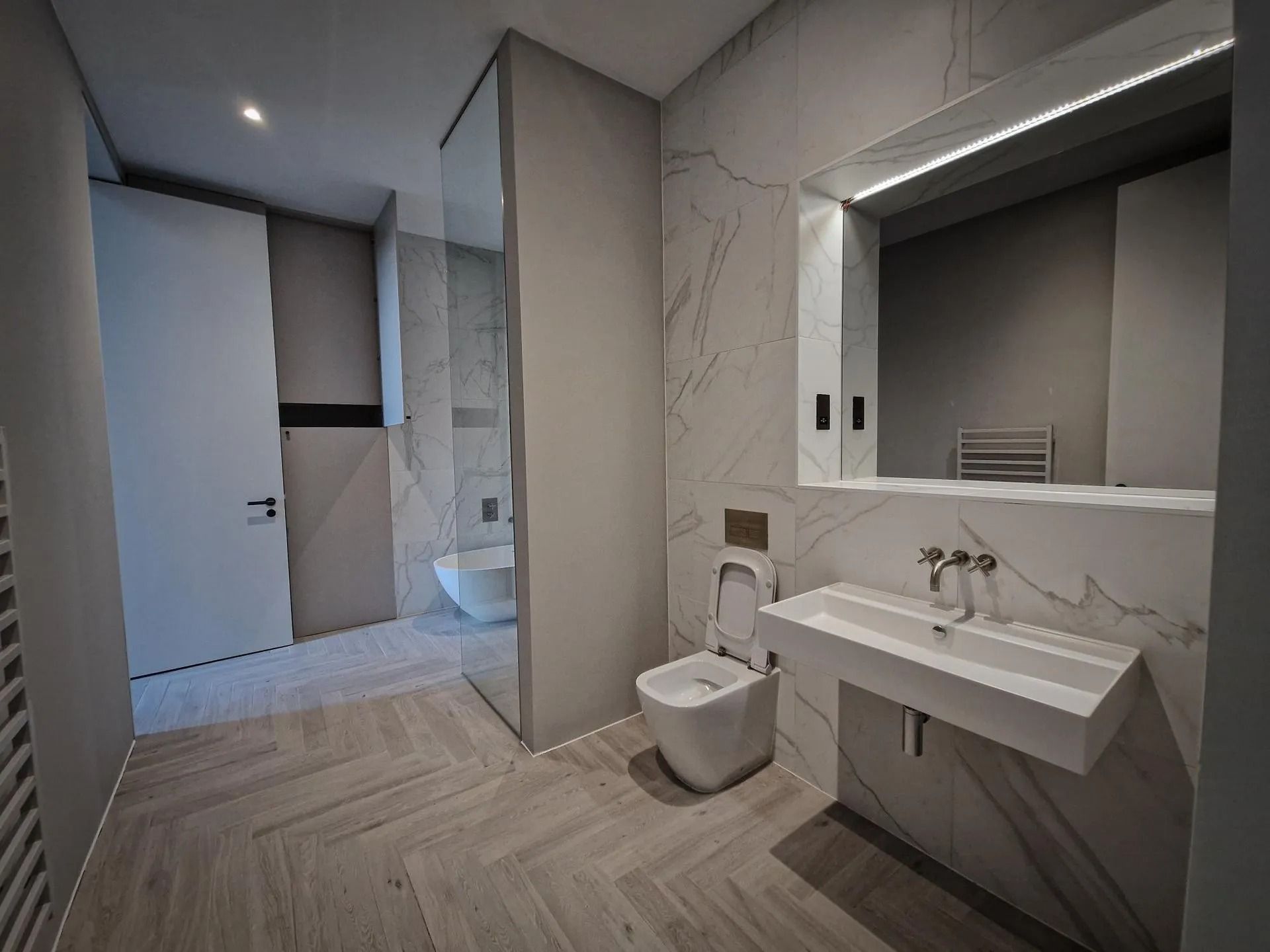 Modern bathroom with marble walls, floating sink, toilet, and large mirror; doorway opens to a shower area