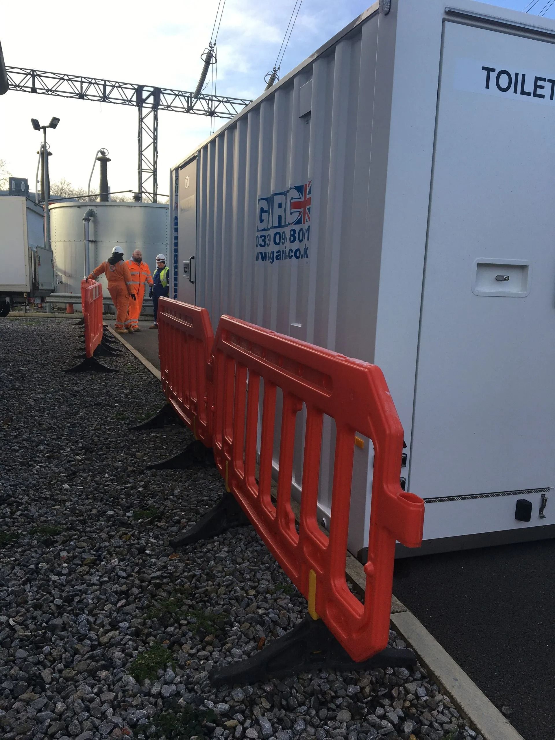 Orange safety barriers beside a white container in a gravel industrial yard with workers in orange coveralls.