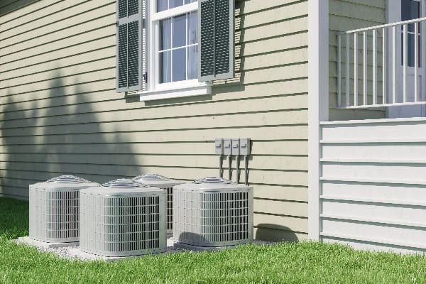 Three white HVAC condenser units sit on a concrete pad outside a light-sided house with a window and porch railing.