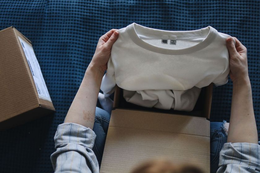 Mani che estraggono una camicia bianca da una scatola di cartone su una superficie di tessuto blu.