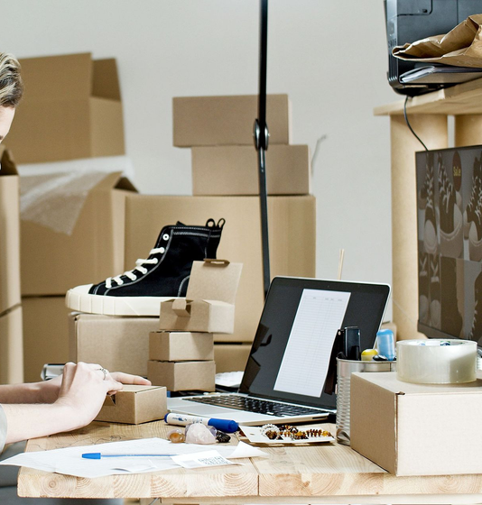 Scatole di scarpe, una scarpa da ginnastica alta nera e materiale da imballaggio su una scrivania in una stanza disordinata.