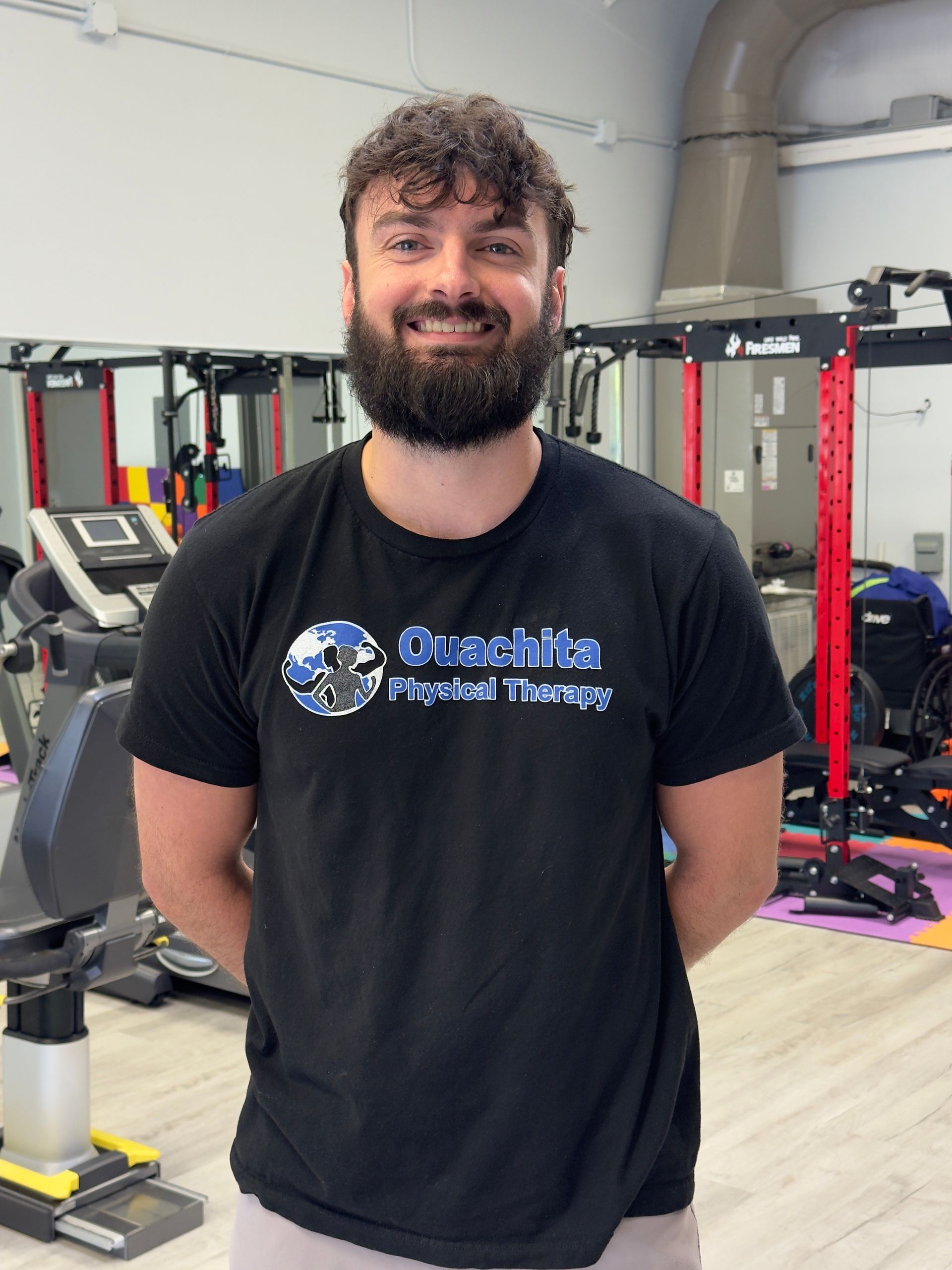 Smiling man in a black T-shirt standing in a gym with weightlifting equipment and exercise machines.