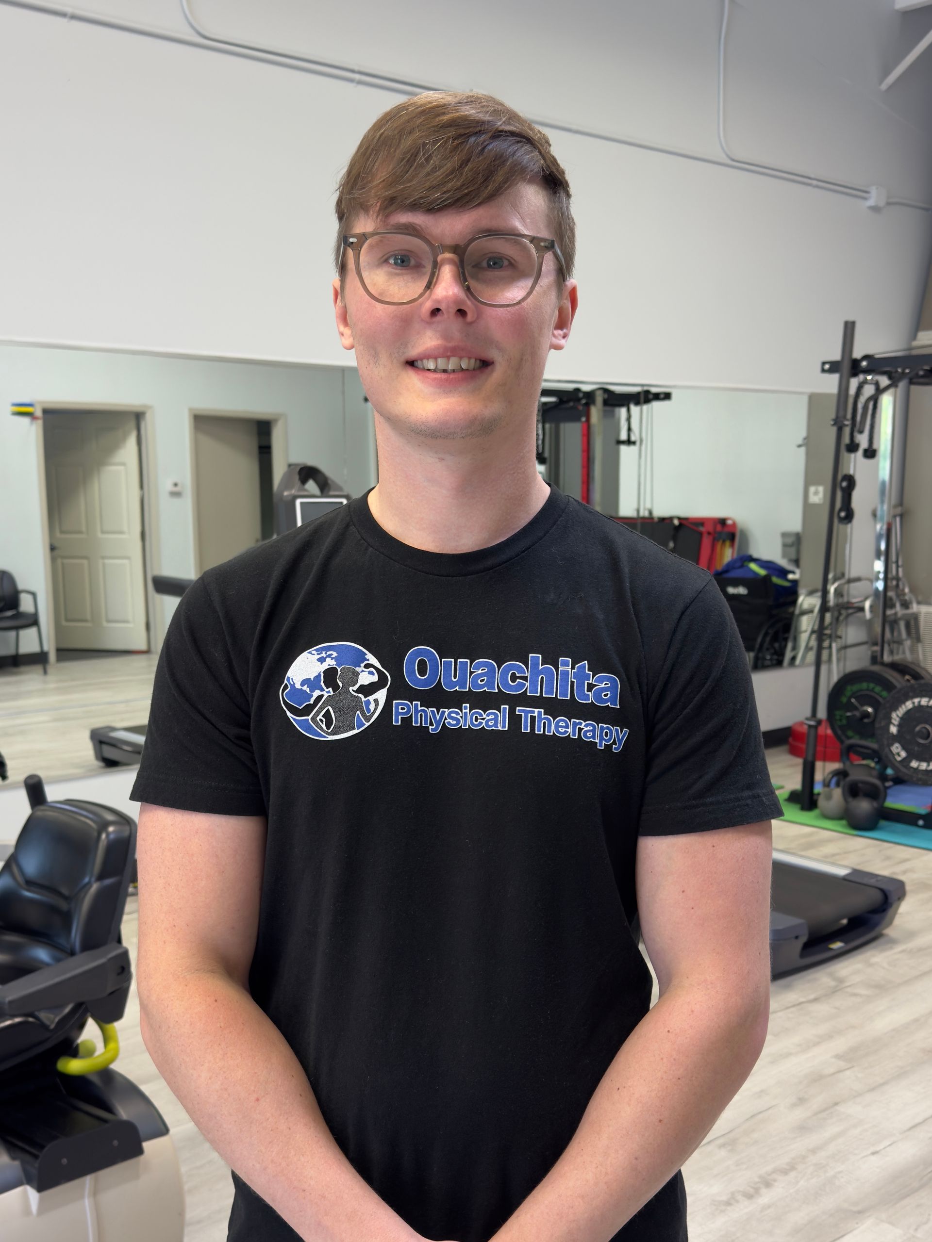 Person standing in a gym, wearing glasses and a black Ouachita Physical Therapy T-shirt, with exercise equipment behind.