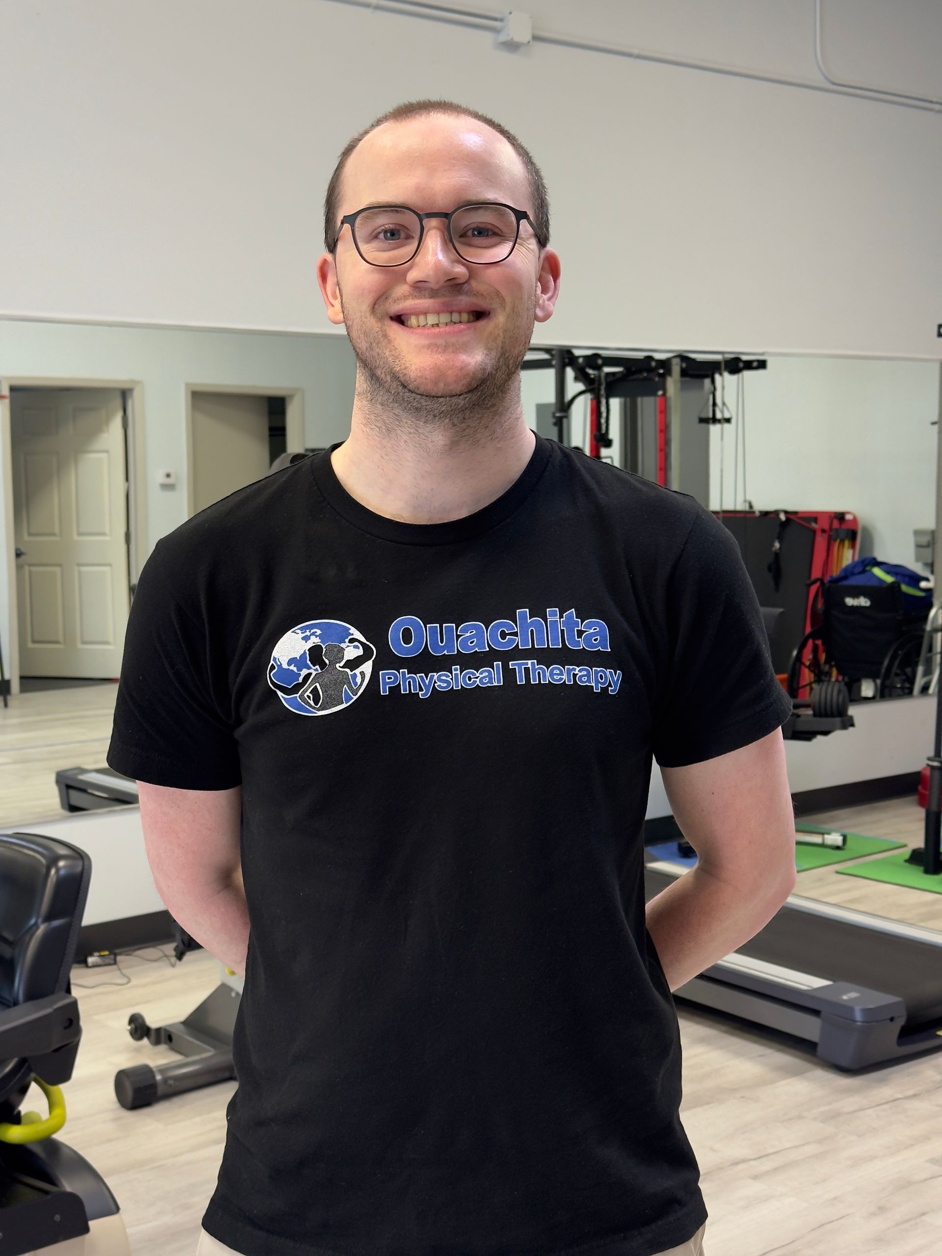 Smiling man in glasses wearing a black T-shirt stands in a gym with exercise equipment behind him