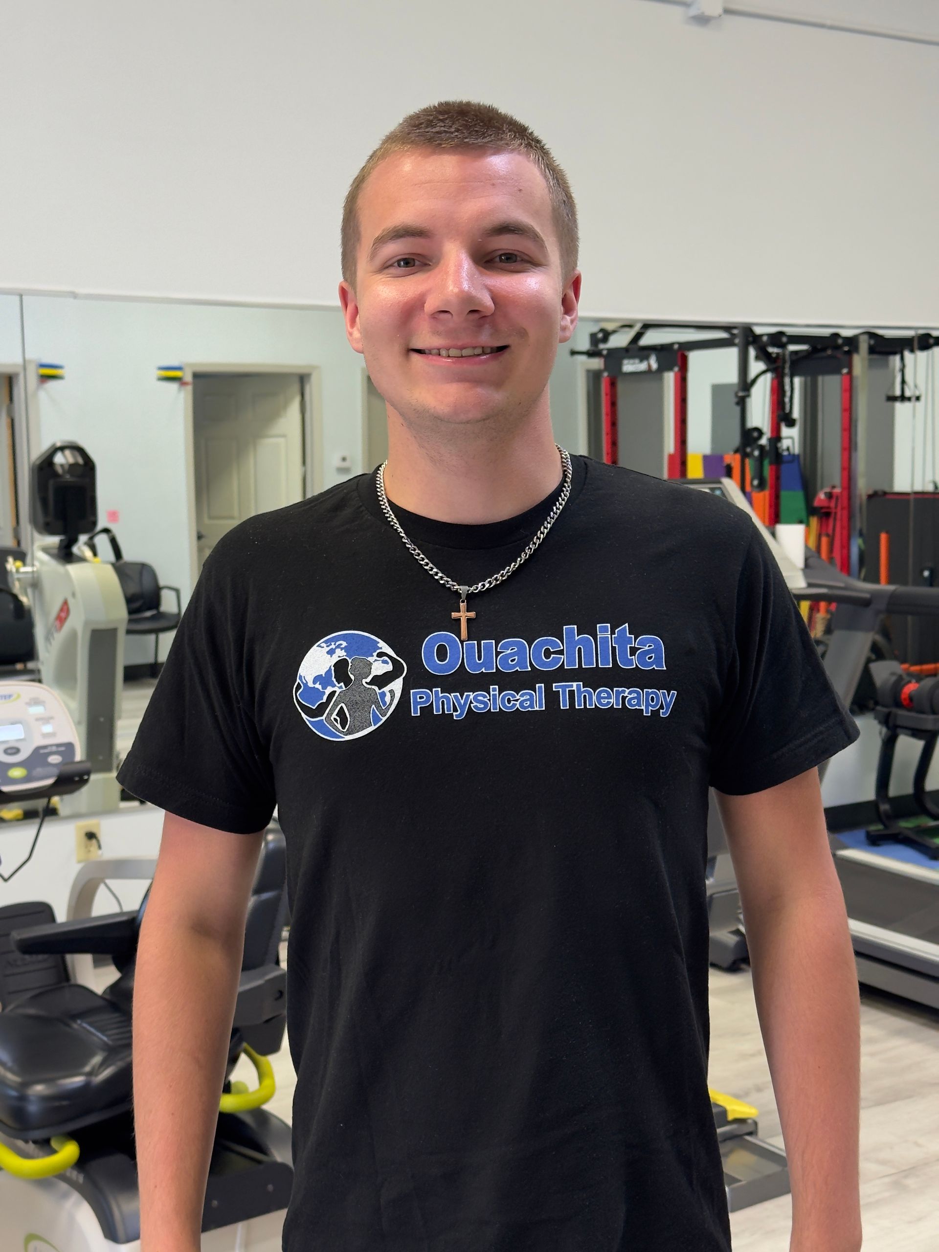 Smiling person in a black T-shirt standing in a gym with exercise equipment in the background