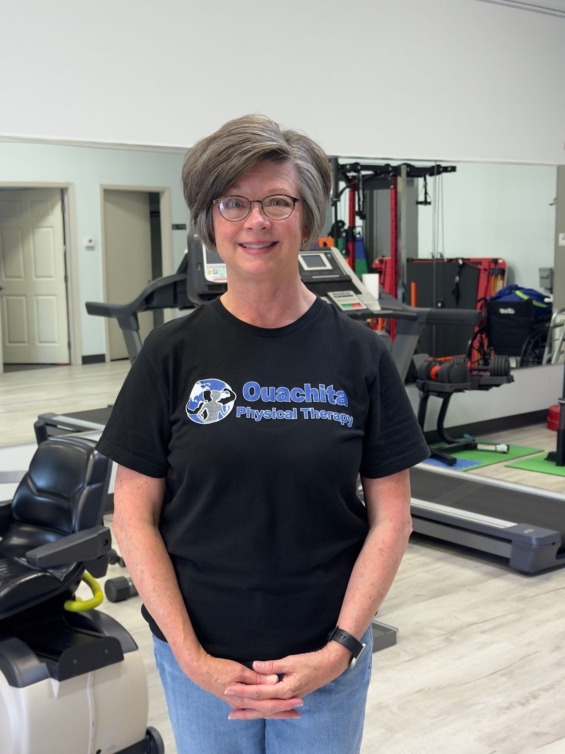 Woman smiling in a gym, wearing a black T-shirt and standing with hands clasped.