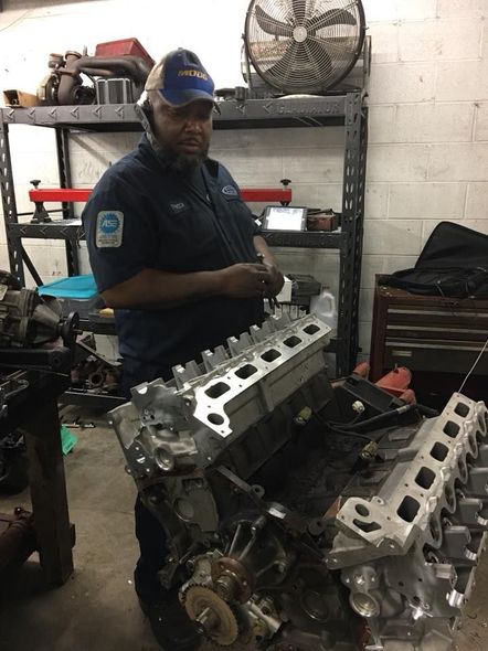 Mechanic holding engine head in a garage; wears blue work uniform and cap. A dark-skinned man; the setting is cluttered.