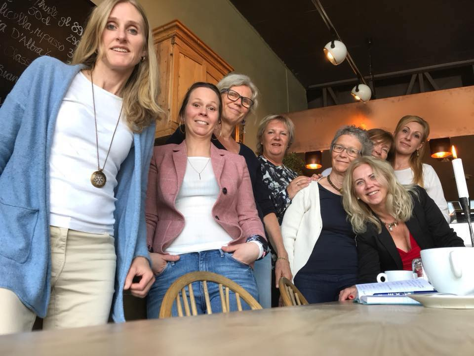 En gruppe smilende mennesker samlet seg innendørs på en kafé, lent rundt et bord.