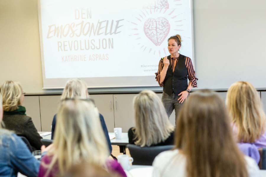 Foredragsholder som presenterer på et klasseromsseminar for et oppmerksomt publikum, med et projisert lysbilde bak seg.