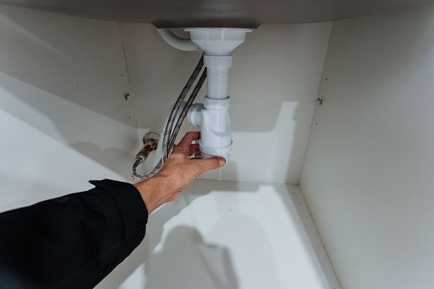 Hand reaching under a sink to adjust the white drain pipe and flexible hose. Hand reaching under a sink to adjust the white drain pipe and flexible hose.