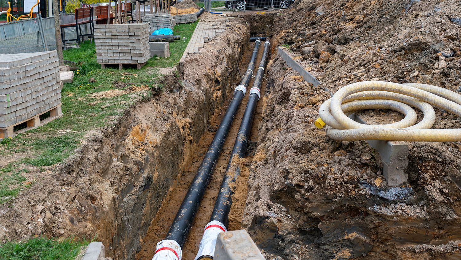 Trenched utility pipes laid in a dirt construction site beside coiled hose and concrete blocks. Trenched utility pipes laid in a dirt construction site beside coiled hose and concrete blocks.