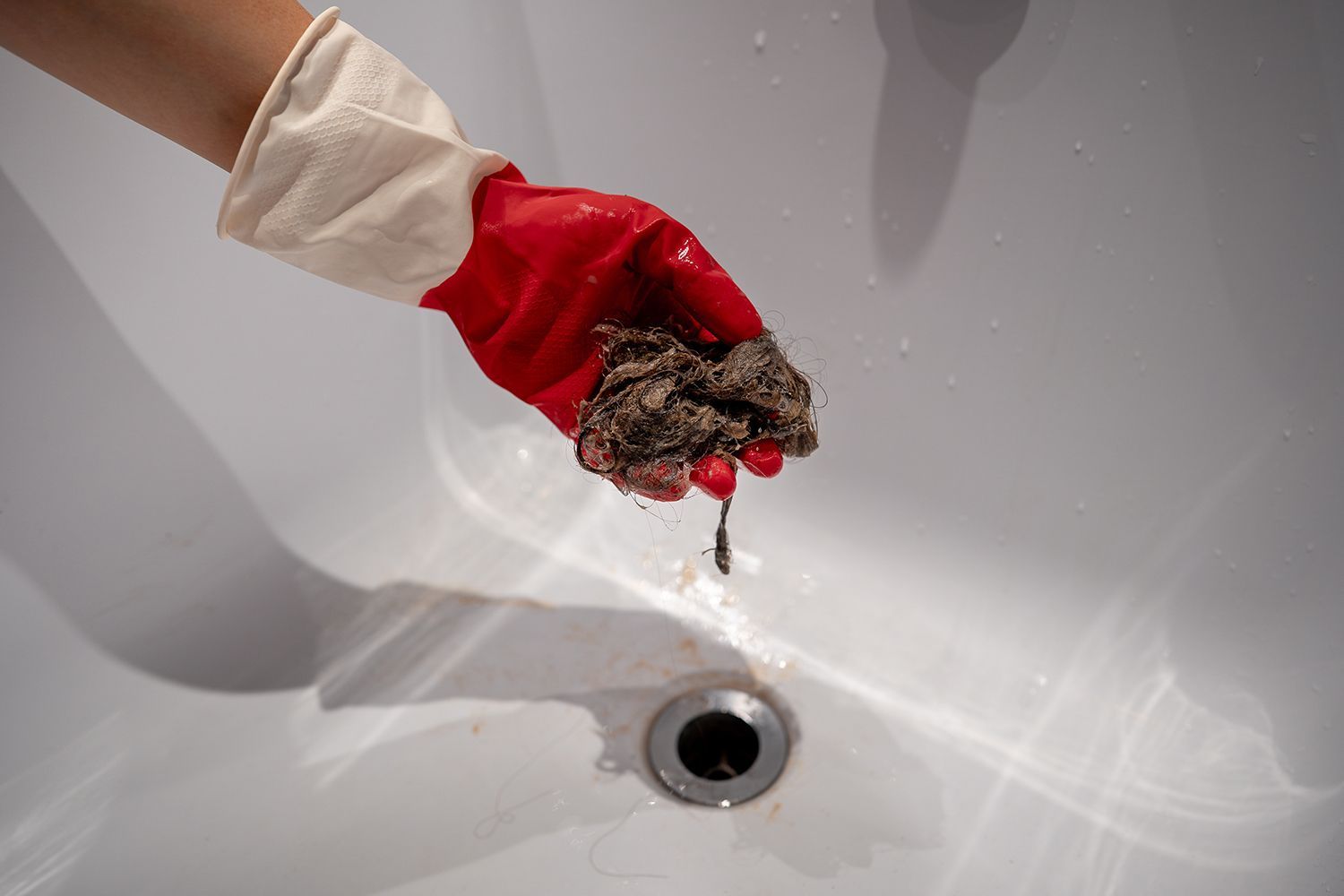 Gloved hand holding clumped debris over a sink drain, with water dripping into the basin Gloved hand holding clumped debris over a sink drain, with water dripping into the basin