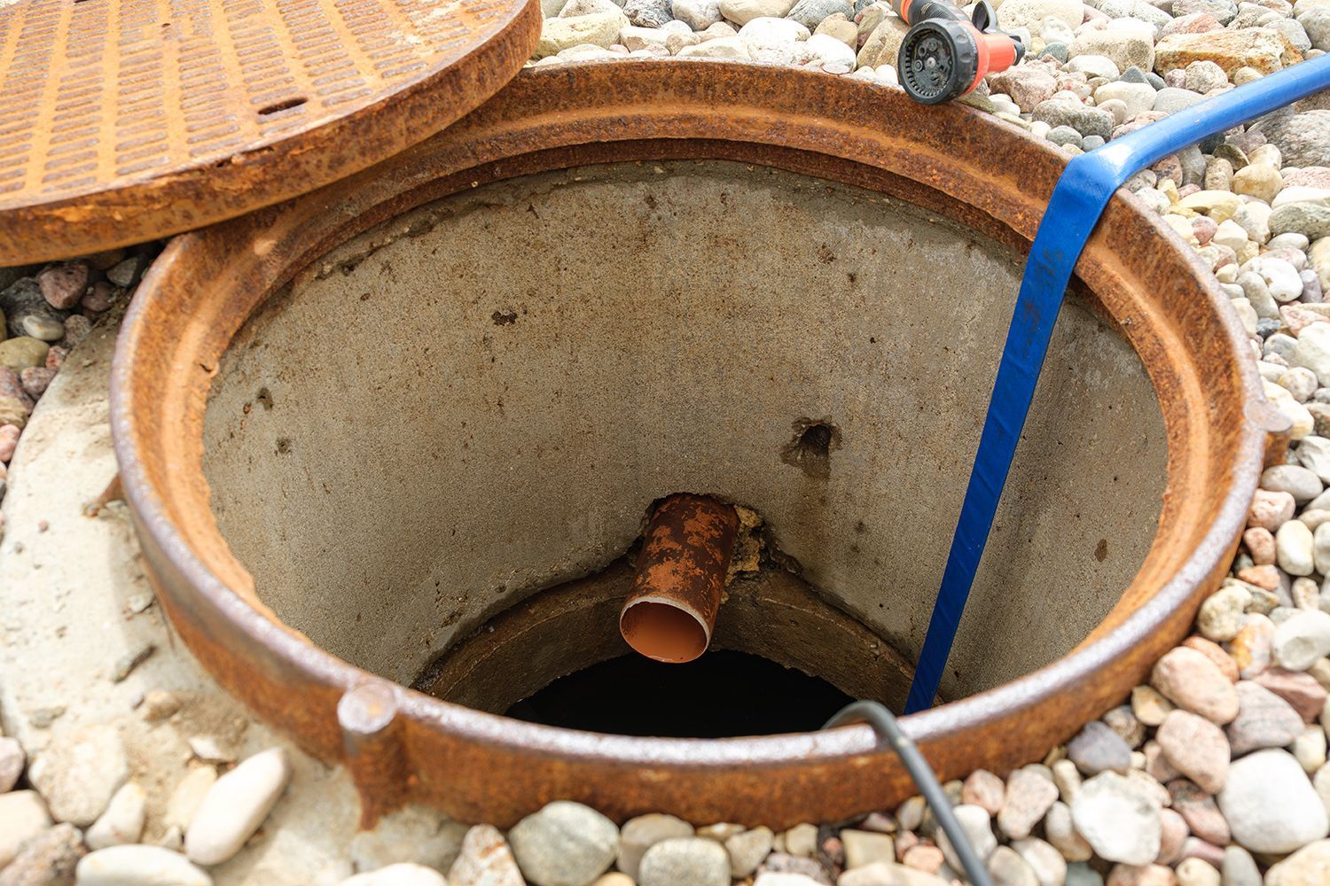Open concrete utility pit with exposed pipe and blue hose among white pebbles Open concrete utility pit with exposed pipe and blue hose among white pebbles