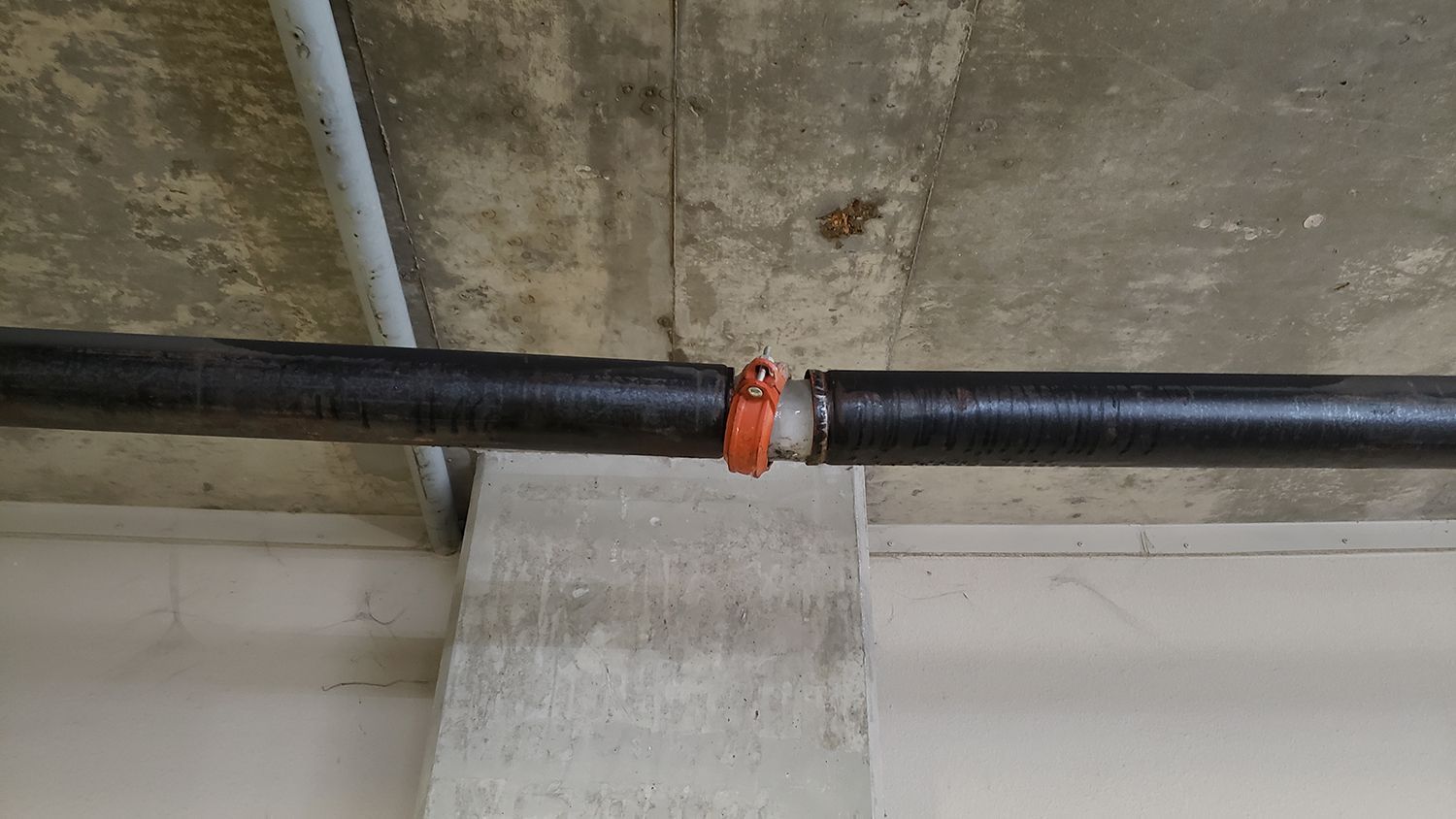 Black pipe with orange clamp running across a concrete ceiling near a wall.