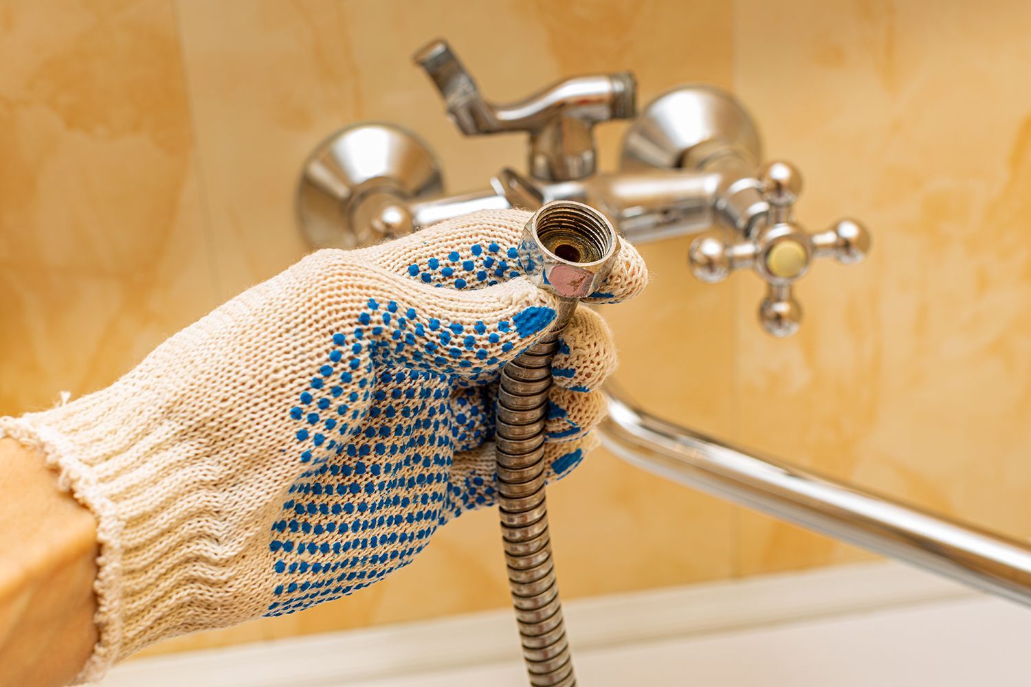 Gloved hand spraying bathroom faucet with a hose nozzle, cleaning chrome fixture and tiled wall
