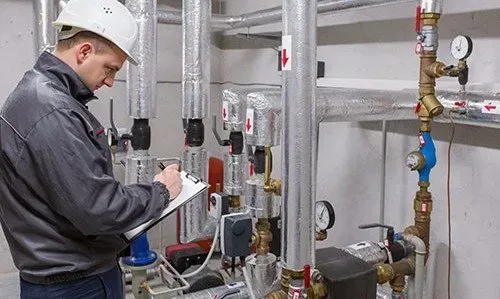 Technician in a gray jacket writes notes beside industrial pipes and gauges in a utility room