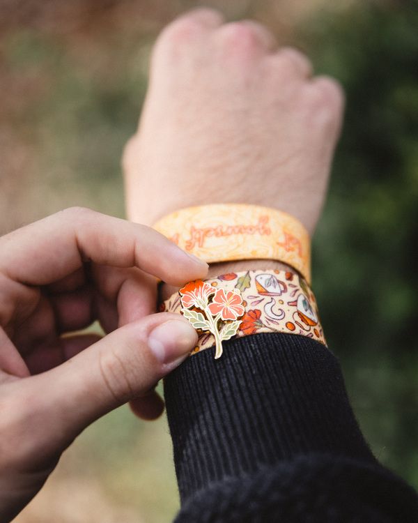 A person's hand adjusts a patterned fabric bracelet with a decorative orange flower charm over a plain yellow wristband.
