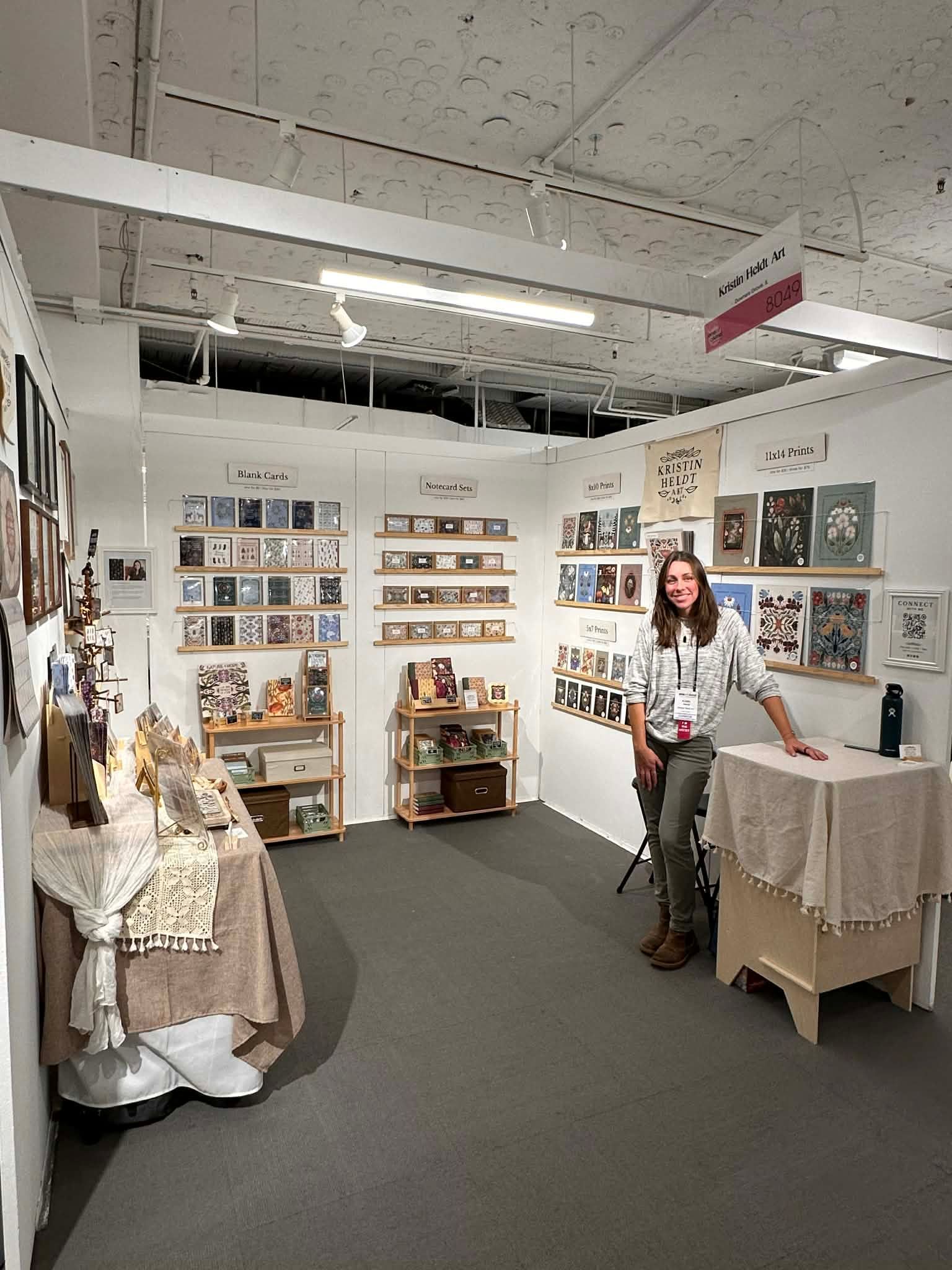 A person stands in a well-lit booth displaying various art prints and books, with shelves and covered tables.