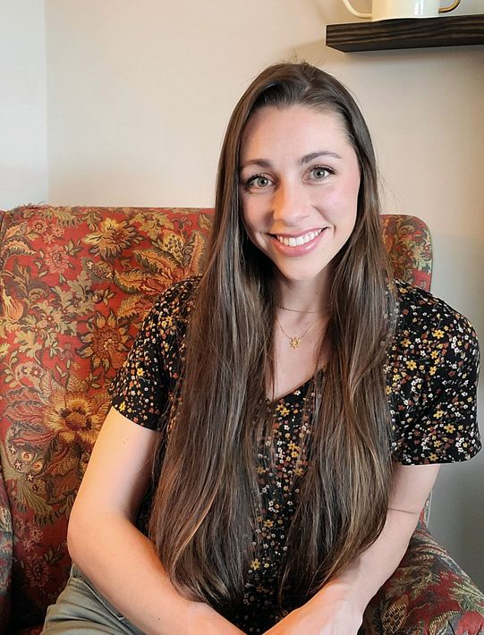 A person with long brown hair, wearing a black floral top, smiles while sitting in a patterned floral armchair.