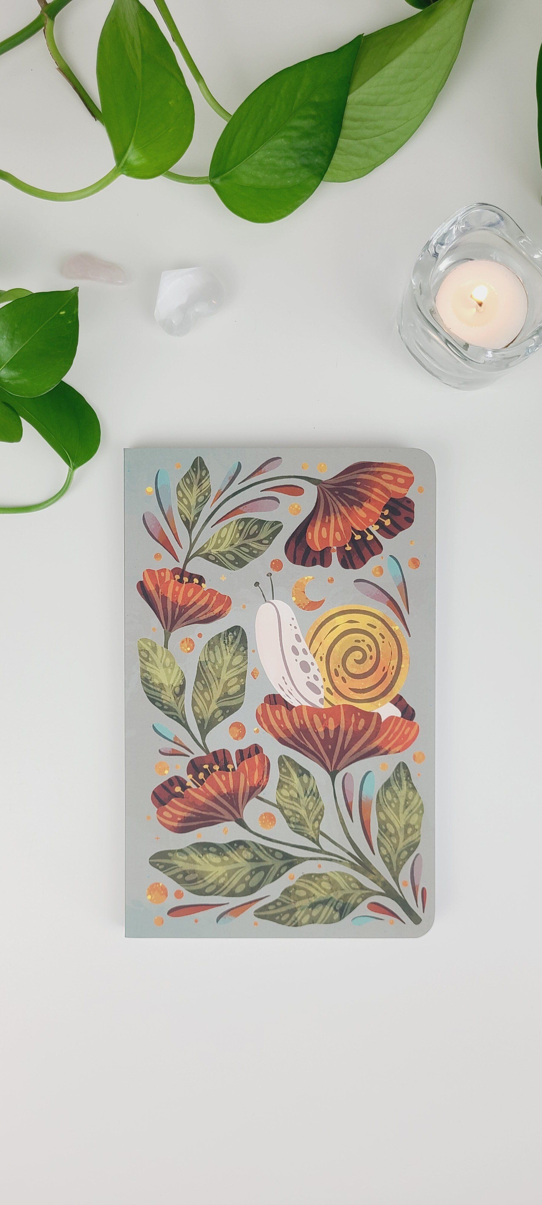 a notebook with a snail on it is sitting on a table next to a plant and a candle .