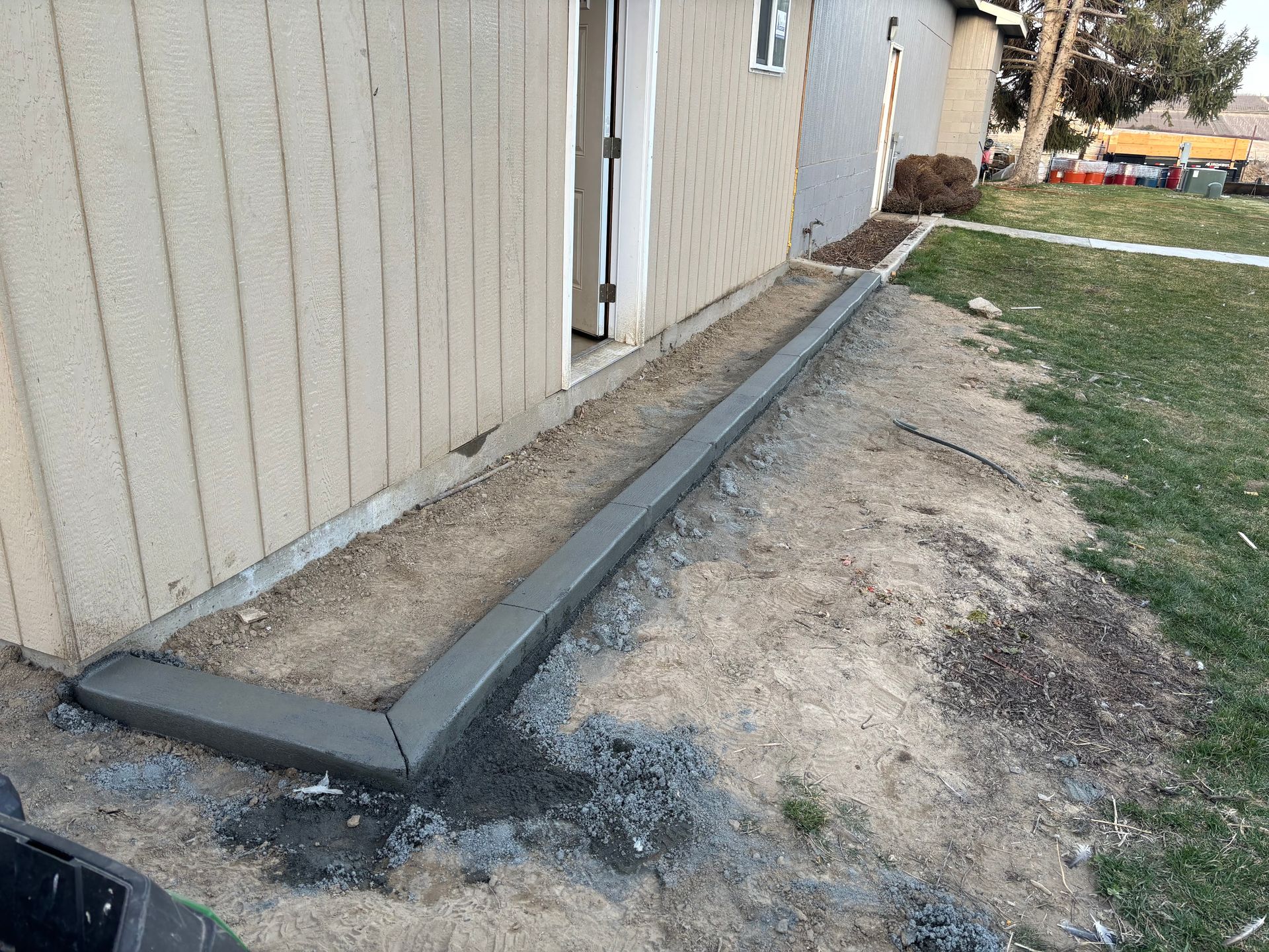 Newly installed concrete landscape edging borders a tan wall and a dirt garden bed alongside a green lawn.