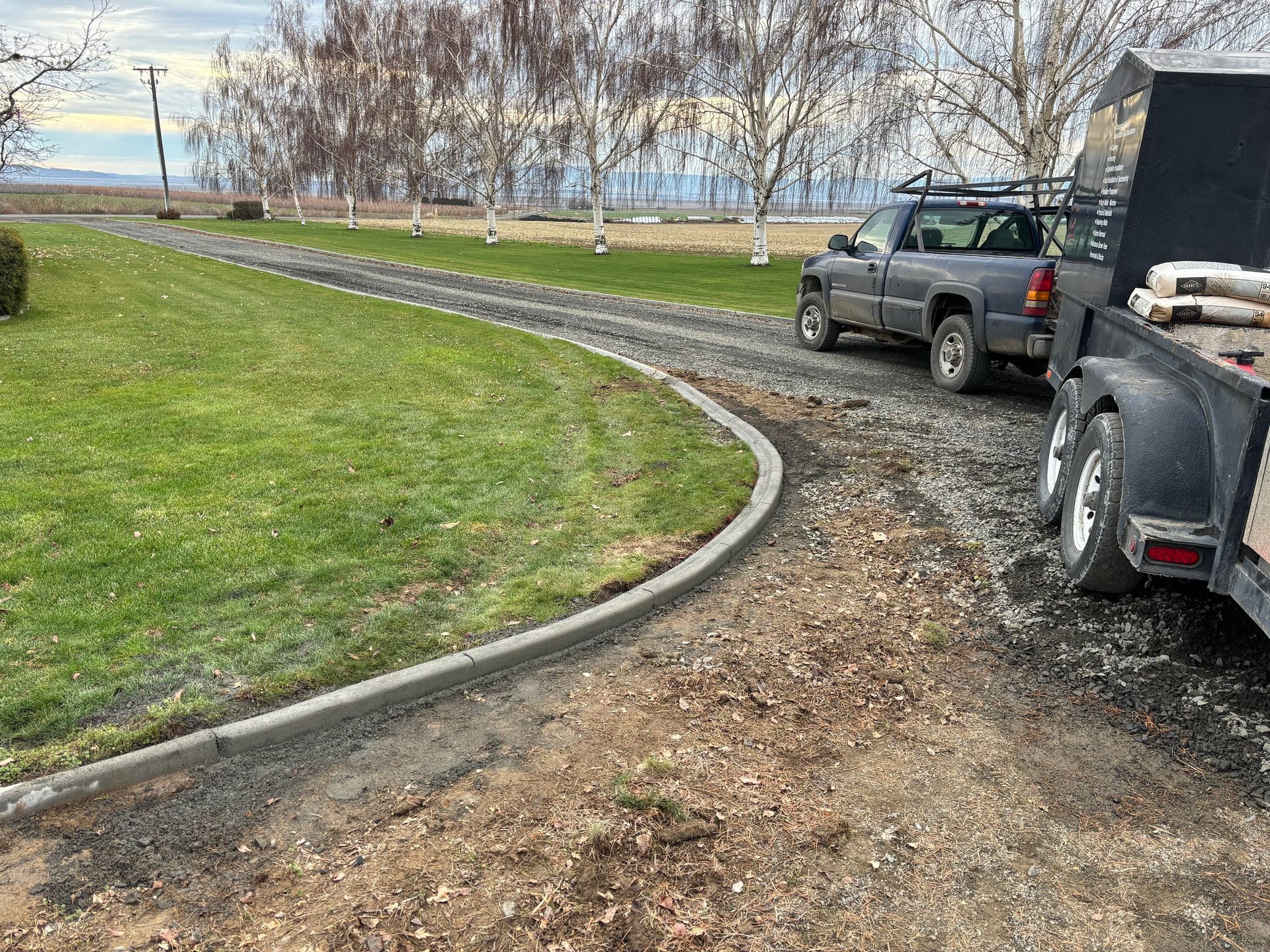 A gray pickup truck towing a utility trailer is parked on a gravel driveway next to a curved concrete curb and lawn.