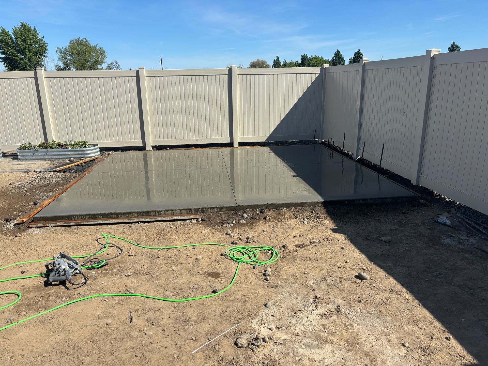 A freshly poured, smooth concrete slab sits in a backyard corner against a light-colored vinyl privacy fence.