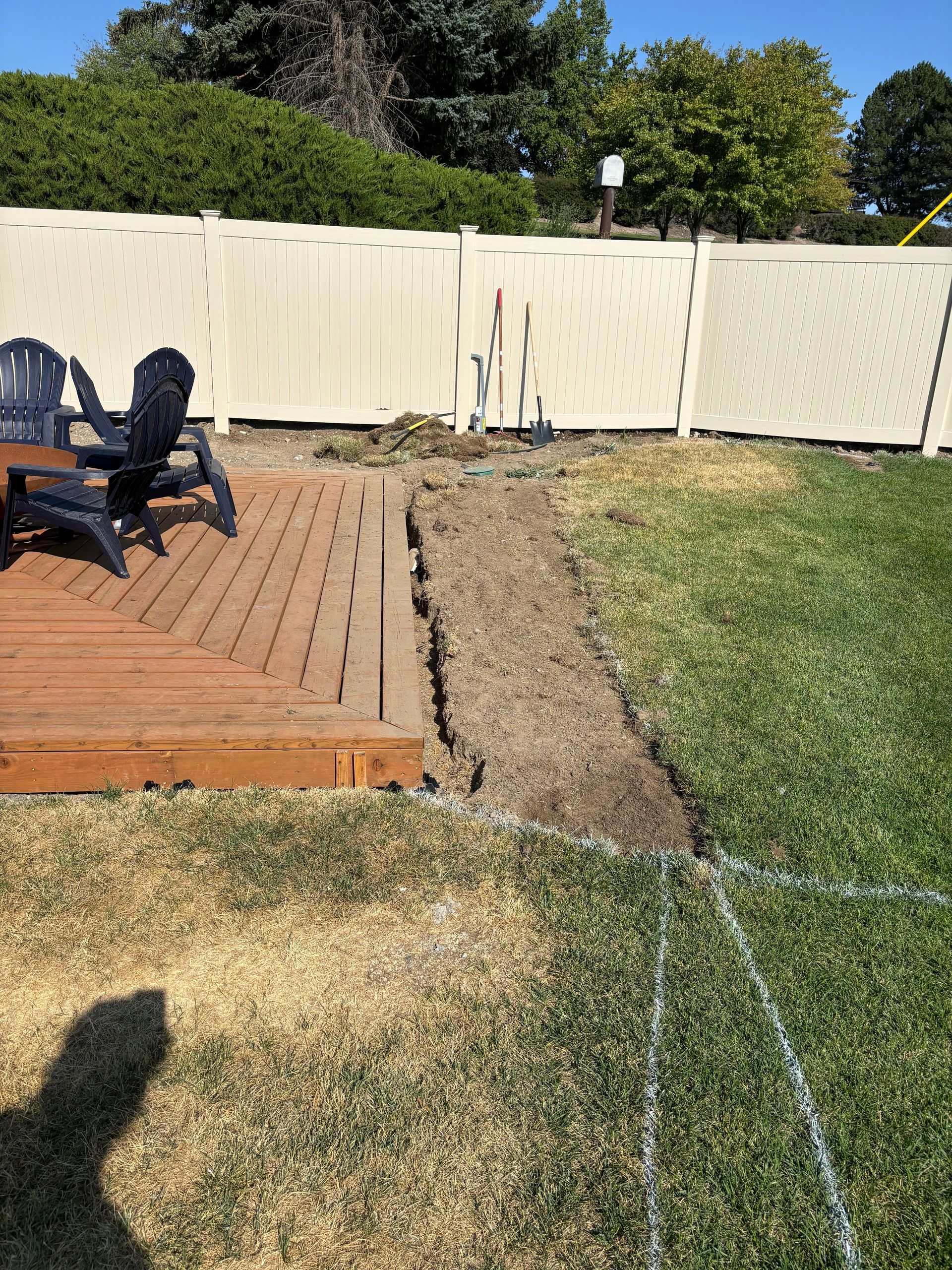 A backyard deck and wooden chairs beside a rectangular patch of turned soil, with a white fence in the background.