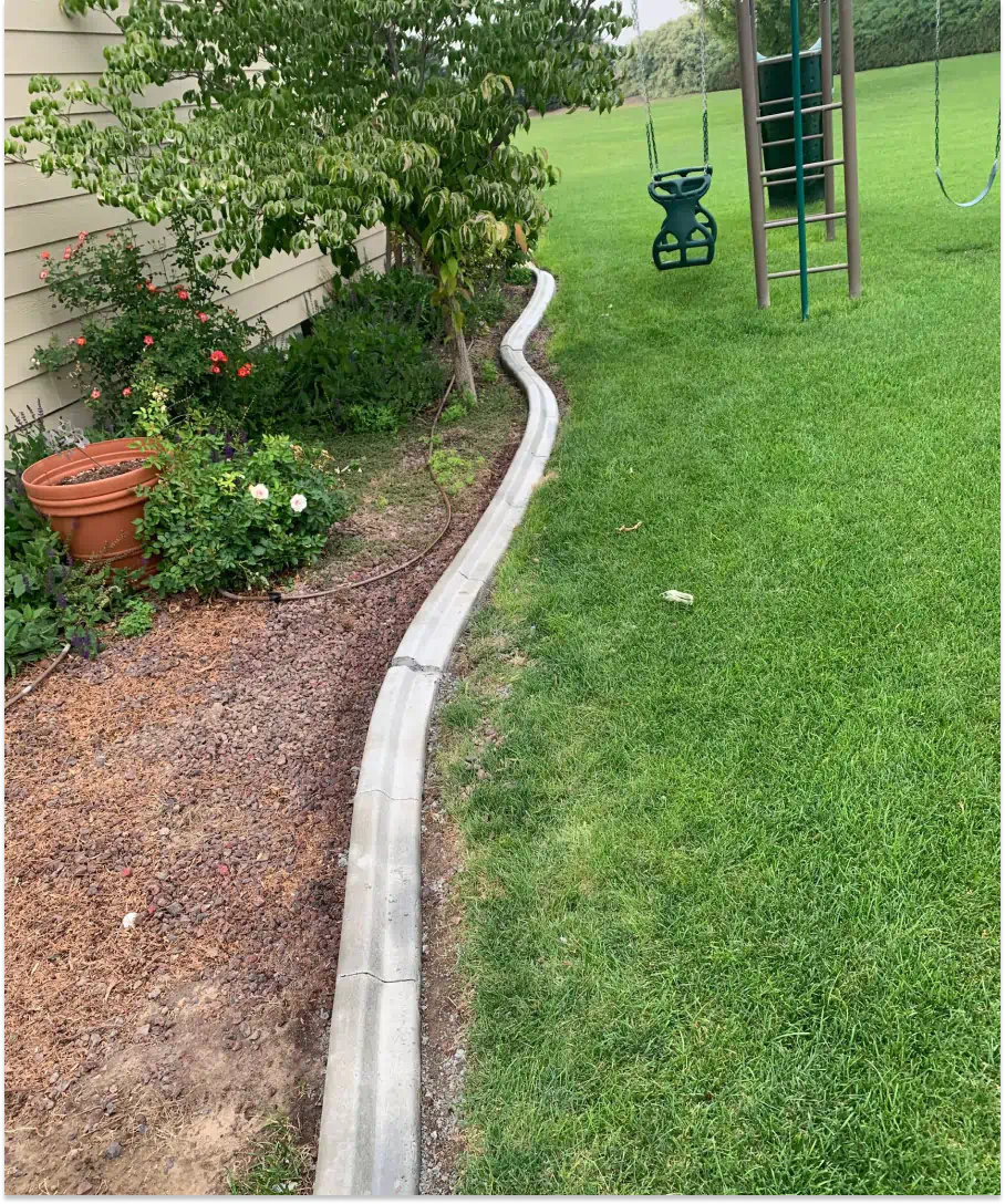 A curved concrete edging separates a mulched garden bed from a grassy lawn next to a house.