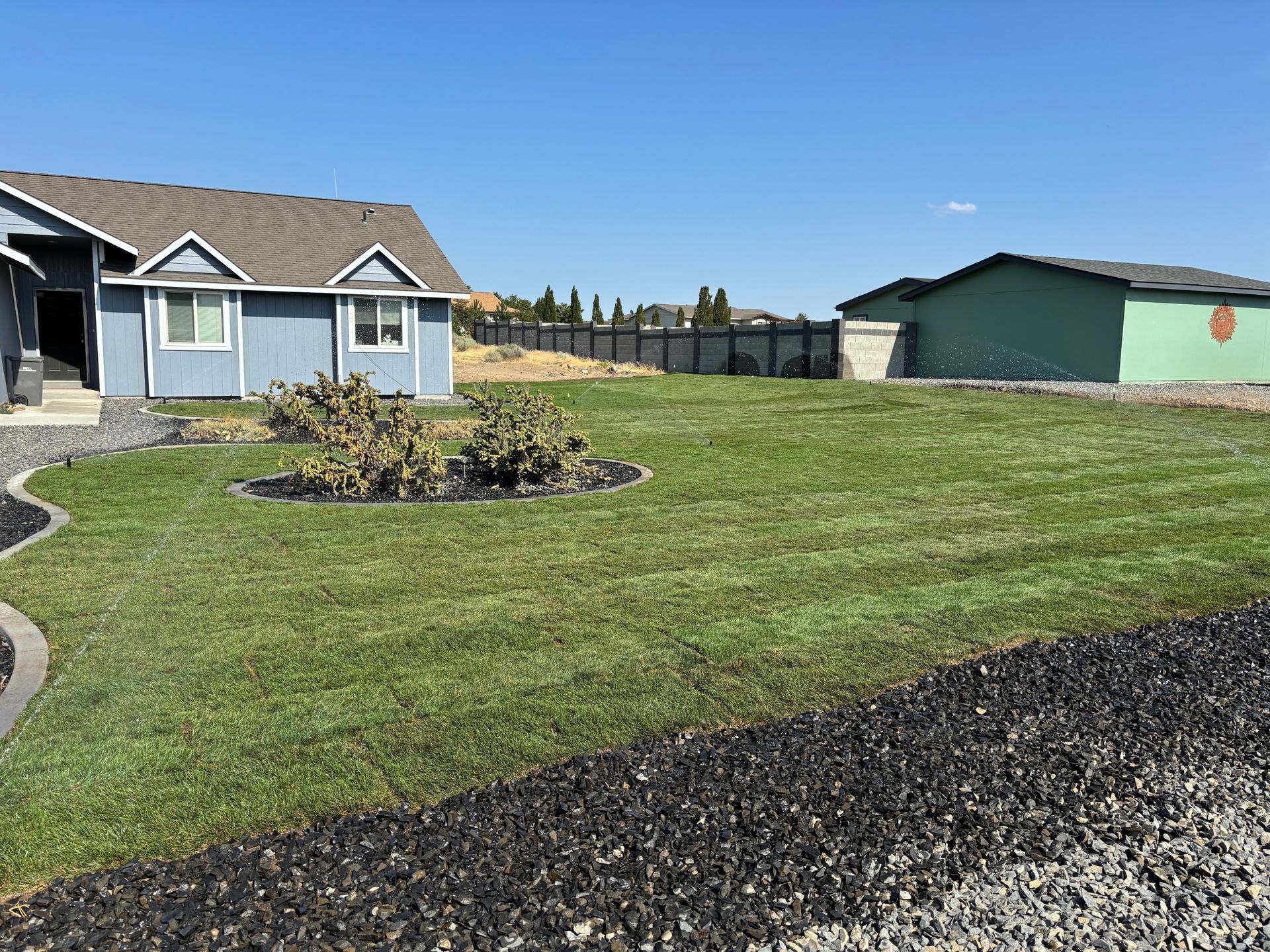 A blue house with a trimmed lawn and a separate green garage under a clear blue sky.