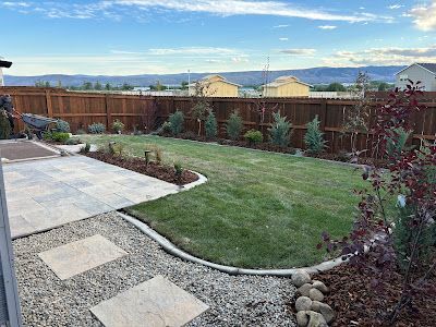 A backyard with a stone patio, gravel pathway, small lawn, and wooden fence, set against a mountain backdrop.