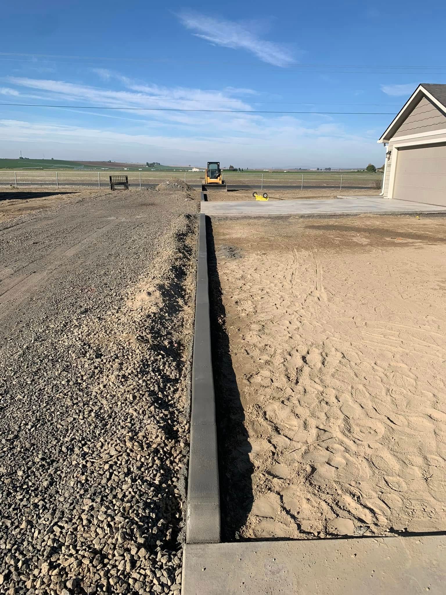 A concrete curb separates a gravel area from a sandy construction site with a house foundation under a blue sky.