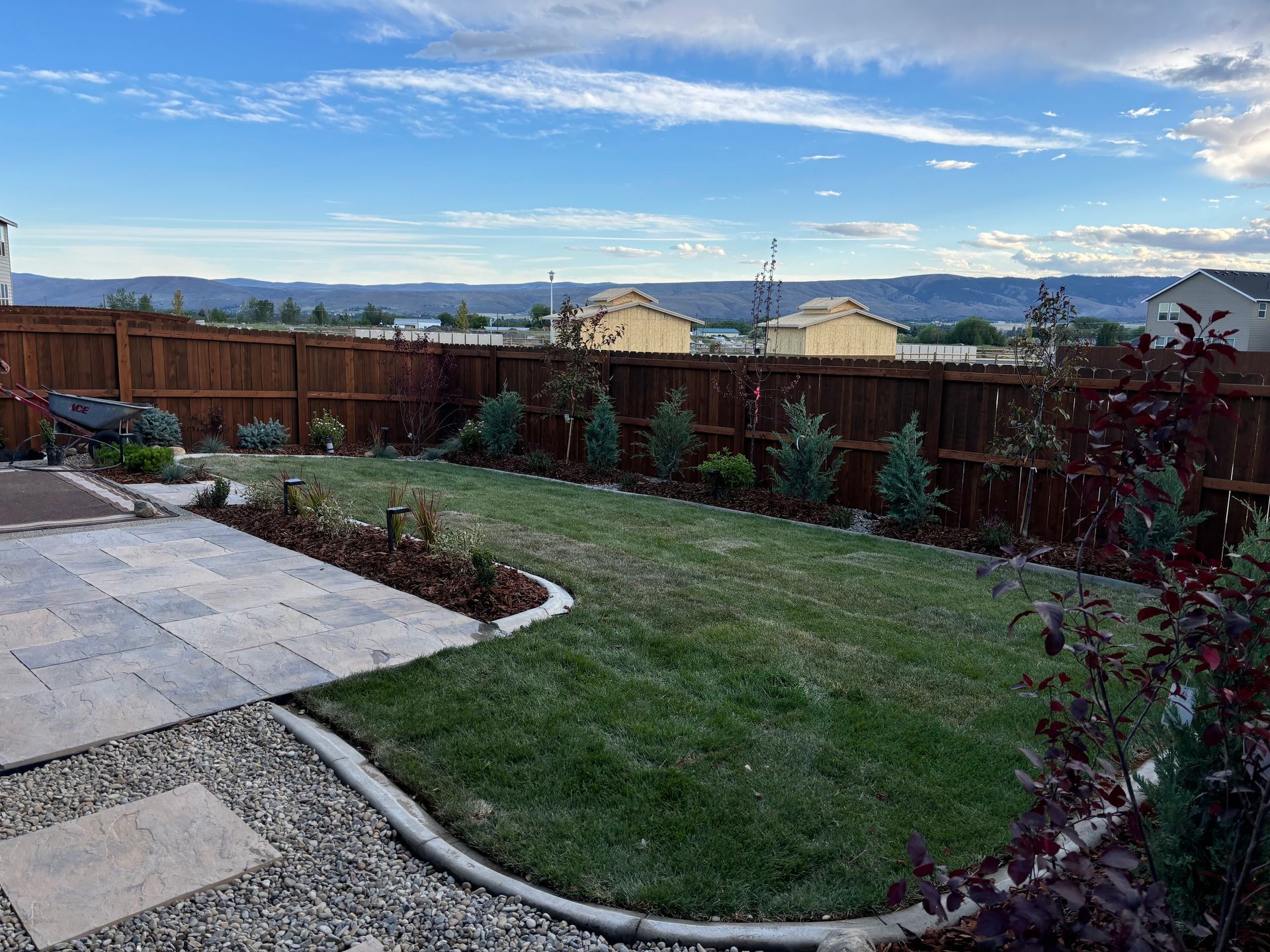 A residential backyard with a paved patio, a grass lawn, a curved concrete border, and a wood fence under a cloudy sky.