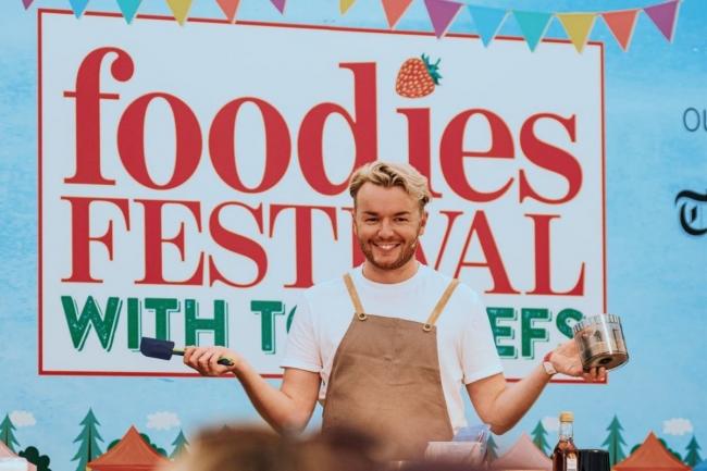 A man stood in front of an audience wearing an apron and holding a spatula.