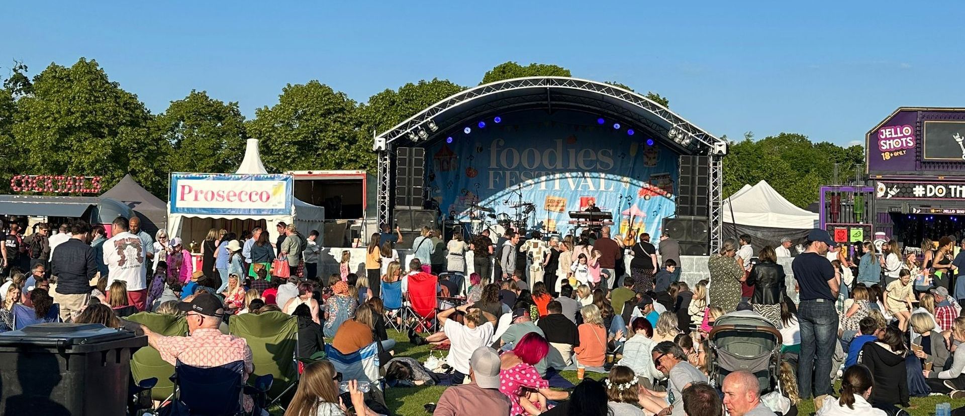 A wide shot of an arc truss stage set up for a live performance, with a large audience in front. There are food and drink stalls either side