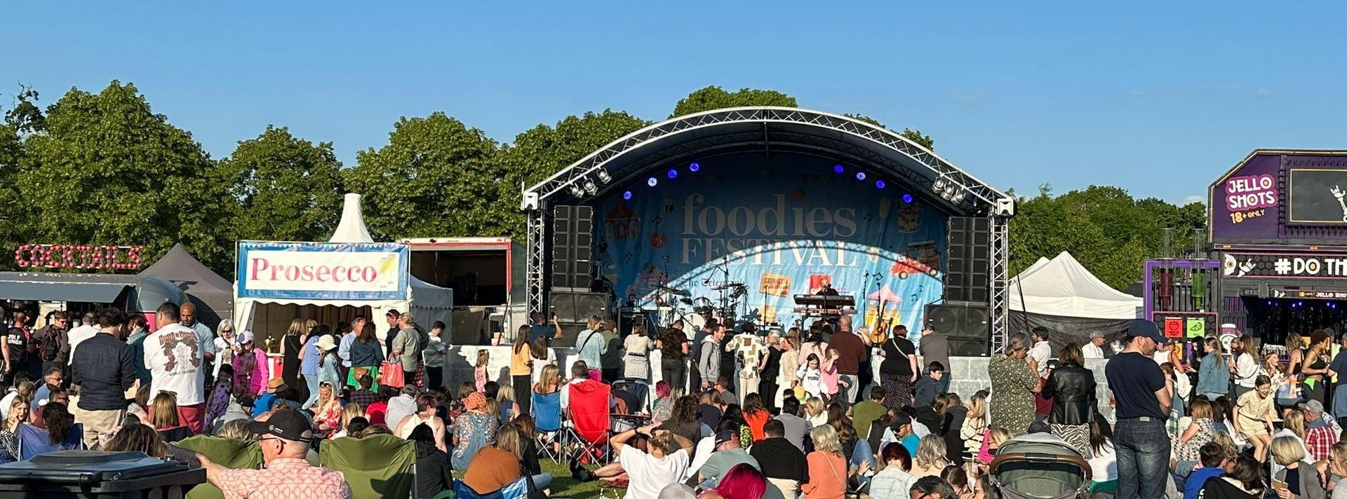 A shot of an arc truss stage in a field with stall holders around it and a large audience in front.